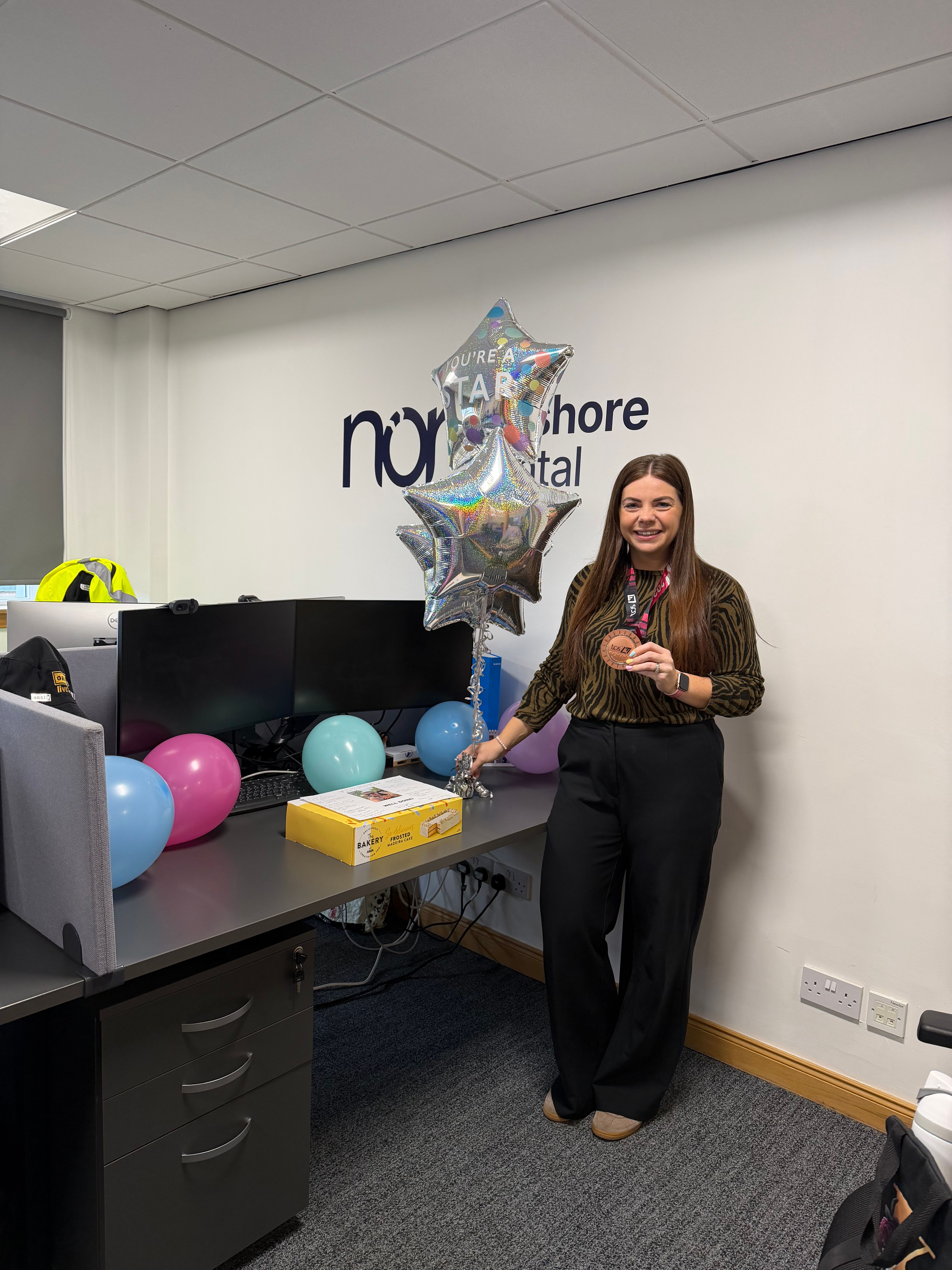 A smiling woman in an office holds a medal, surrounded by "You're a Star" balloons and a gift box, with the "North Shore Capital" logo on the wall.