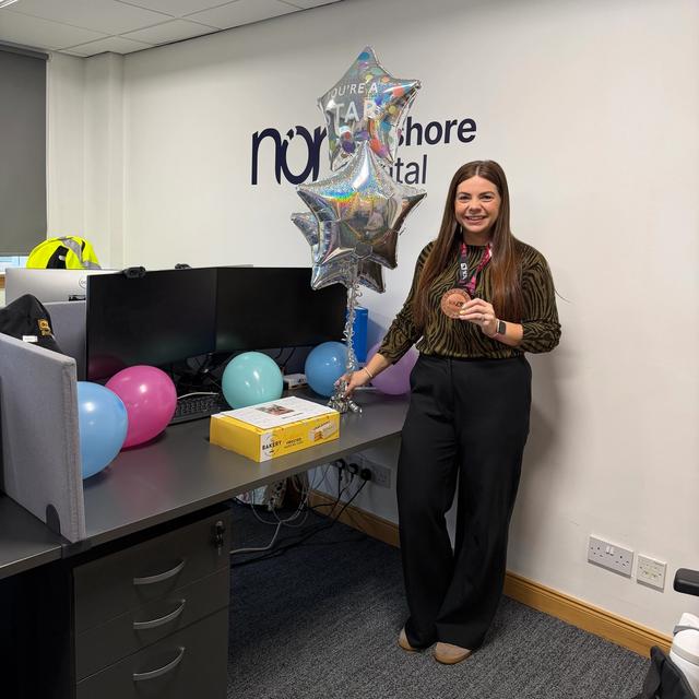 A smiling woman in an office holds a medal, surrounded by "You're a Star" balloons and a gift box, with the "North Shore Capital" logo on the wall.