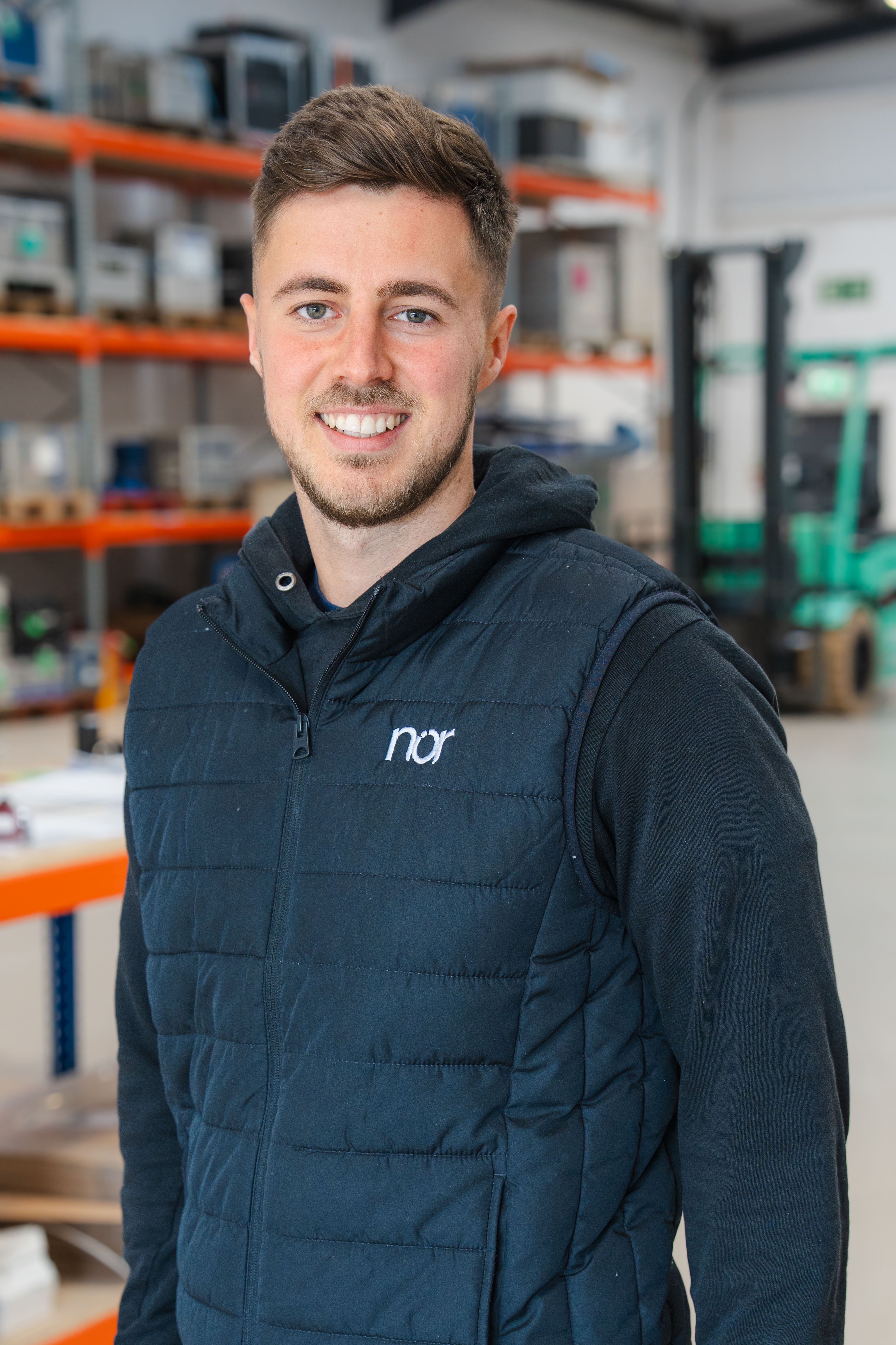 A smiling man wearing a dark blue "nor" vest in a warehouse.