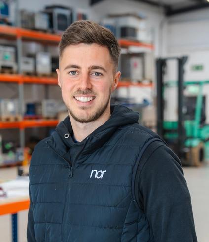 A smiling man wearing a dark blue "nor" vest in a warehouse.