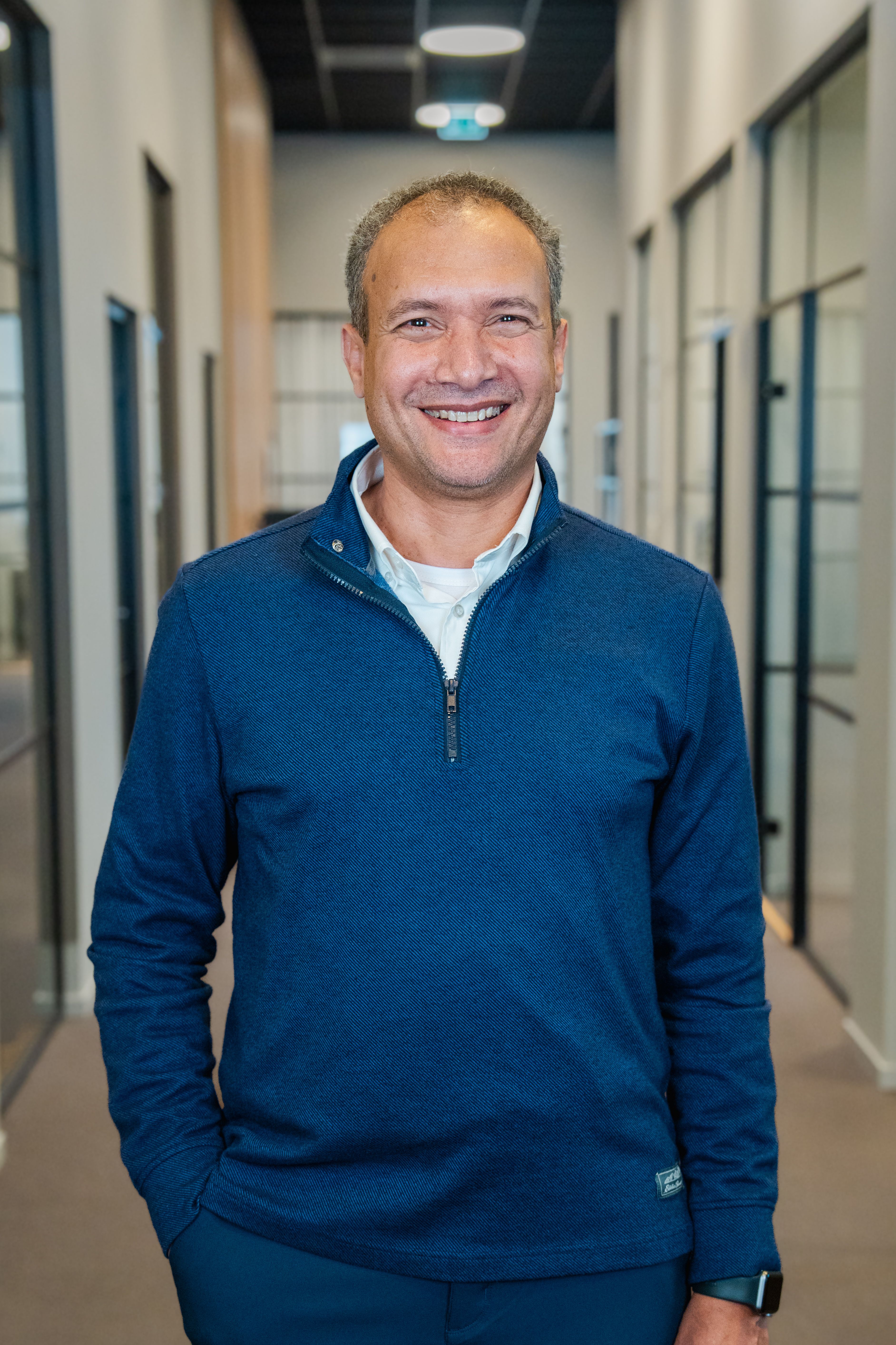 A smiling man wearing a blue quarter-zip shirt stands in a modern office hallway.