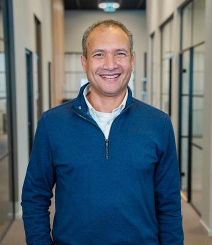 A smiling man wearing a blue quarter-zip shirt stands in a modern office hallway.