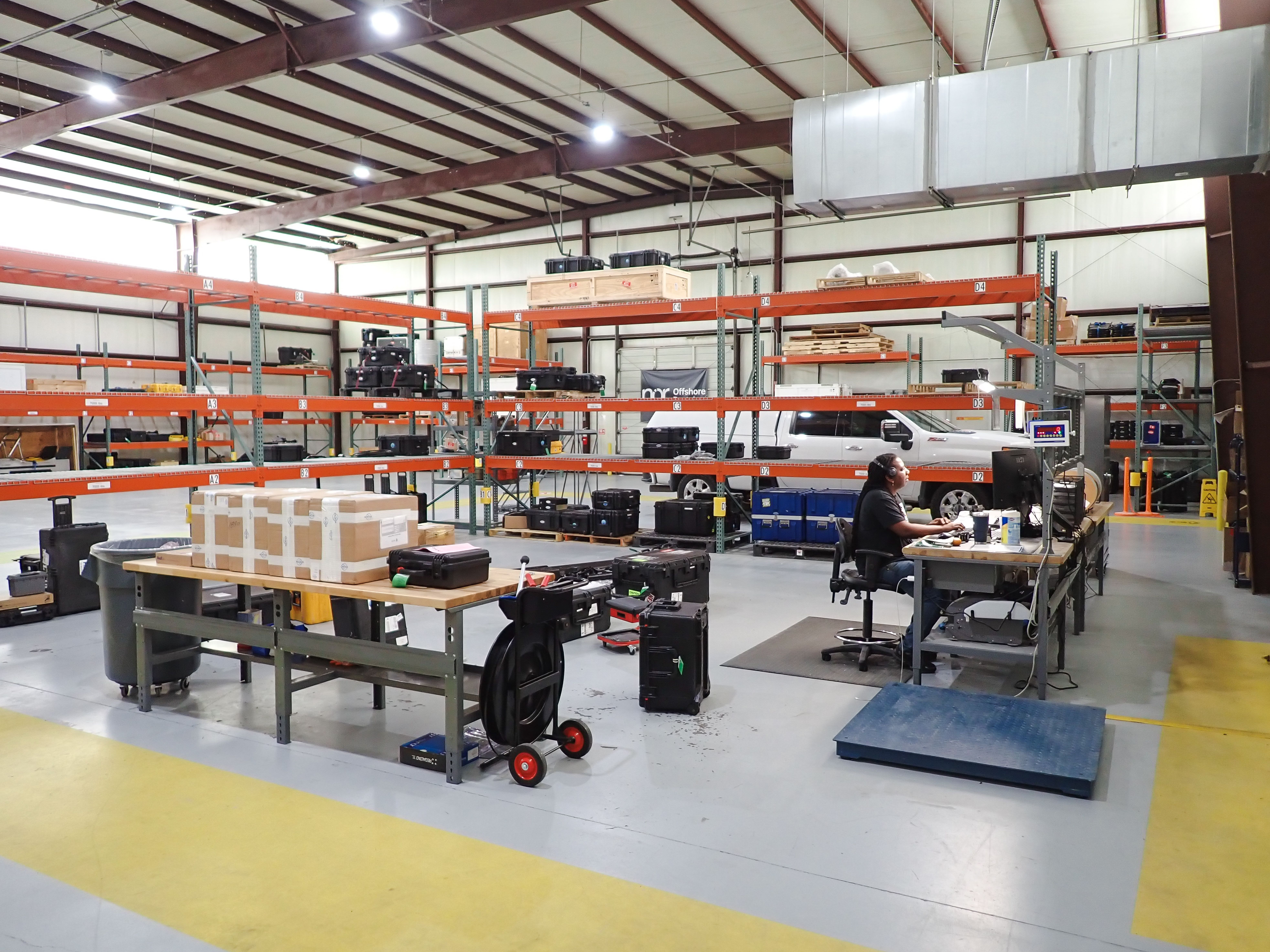 A person works at a desk in a large warehouse filled with equipment on storage racks, with a white pickup truck in the background.