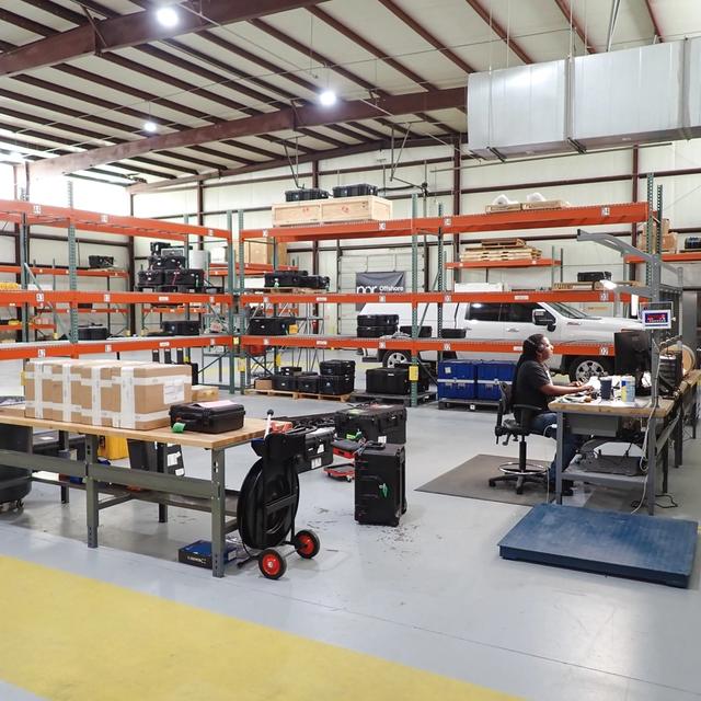 A person works at a desk in a large warehouse filled with equipment on storage racks, with a white pickup truck in the background.