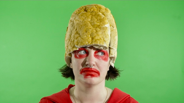 Lara Bell wearing a bread headdress and red face paint against a green background