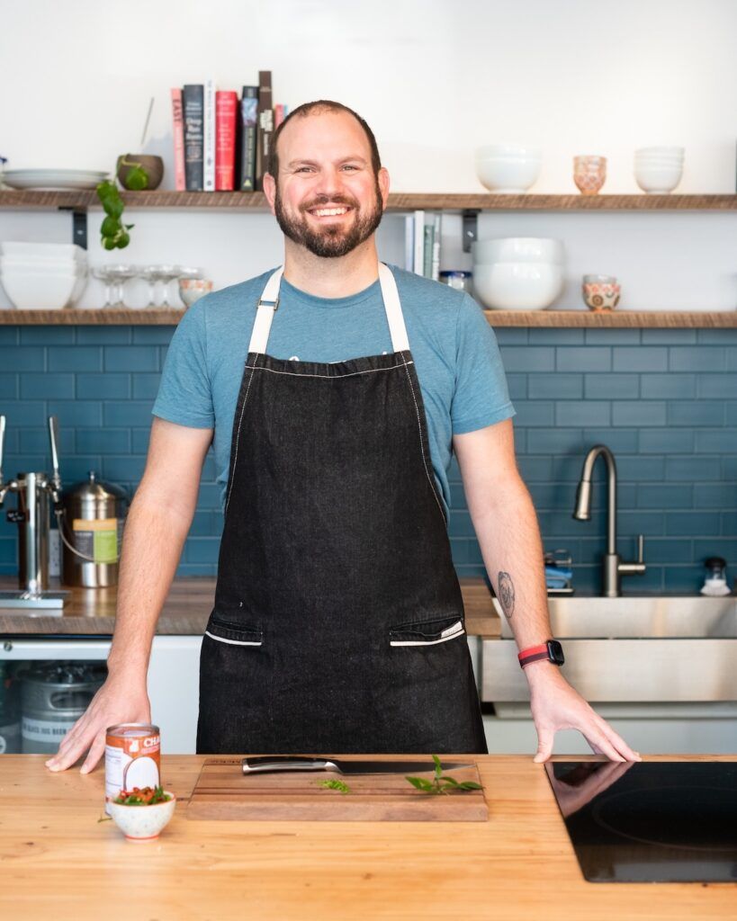 Picture of Nathan in an apron, standing behind a kitchen island (in the Current Wellness kitchen)