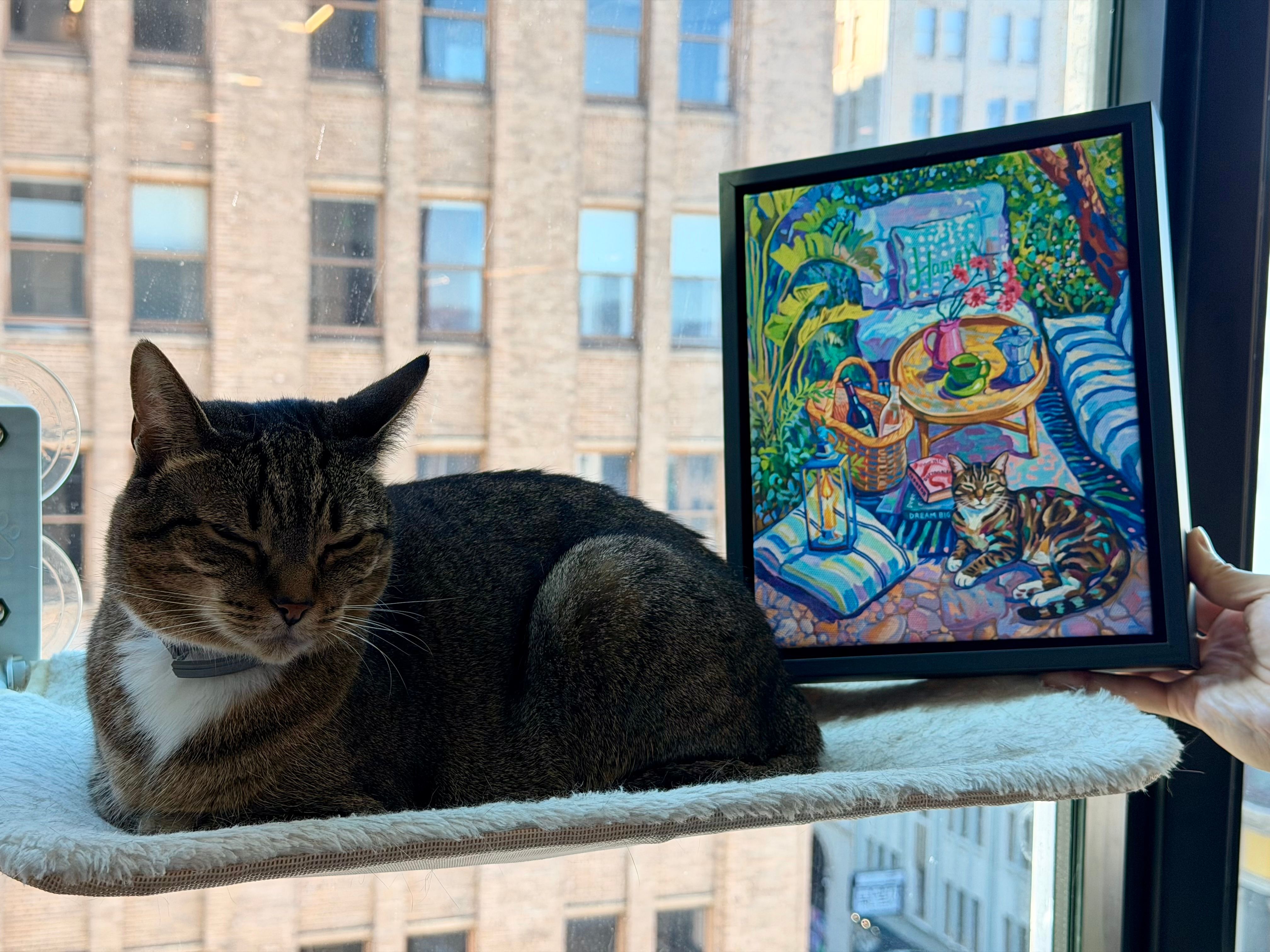 Tabby cat relaxing by a window beside a framed custom cat portrait inspired by a colorful garden scene.