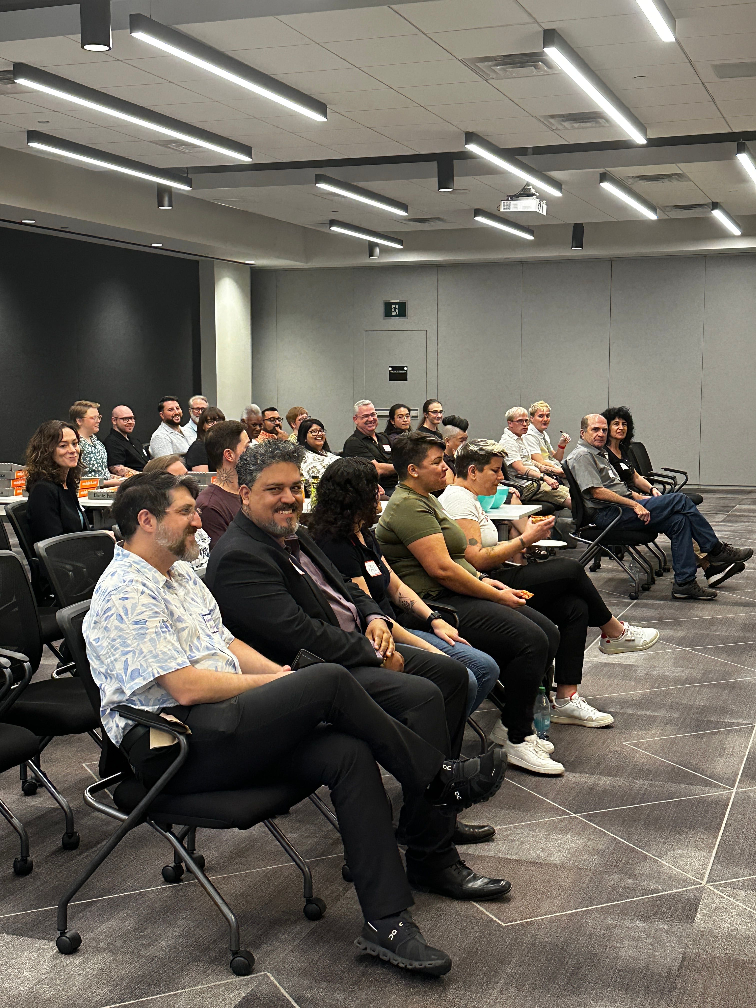 A diverse group of people seated in a conference room, engaged and smiling. The modern setting features rows of chairs and sleek ceiling lights.