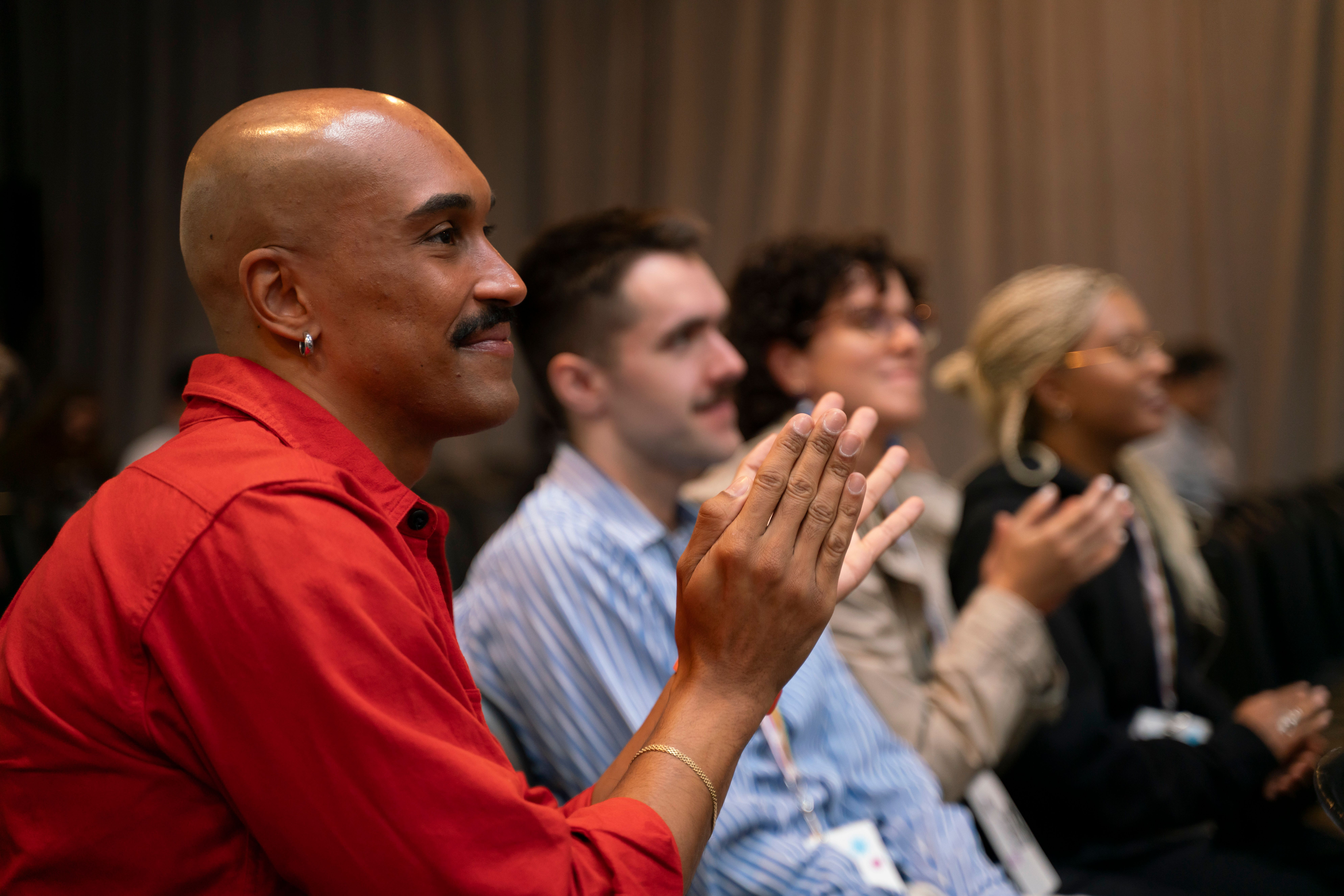 A group of diverse individuals sit in an audience, clapping and smiling. The atmosphere is engaged and positive, with focus on a person in a red shirt.