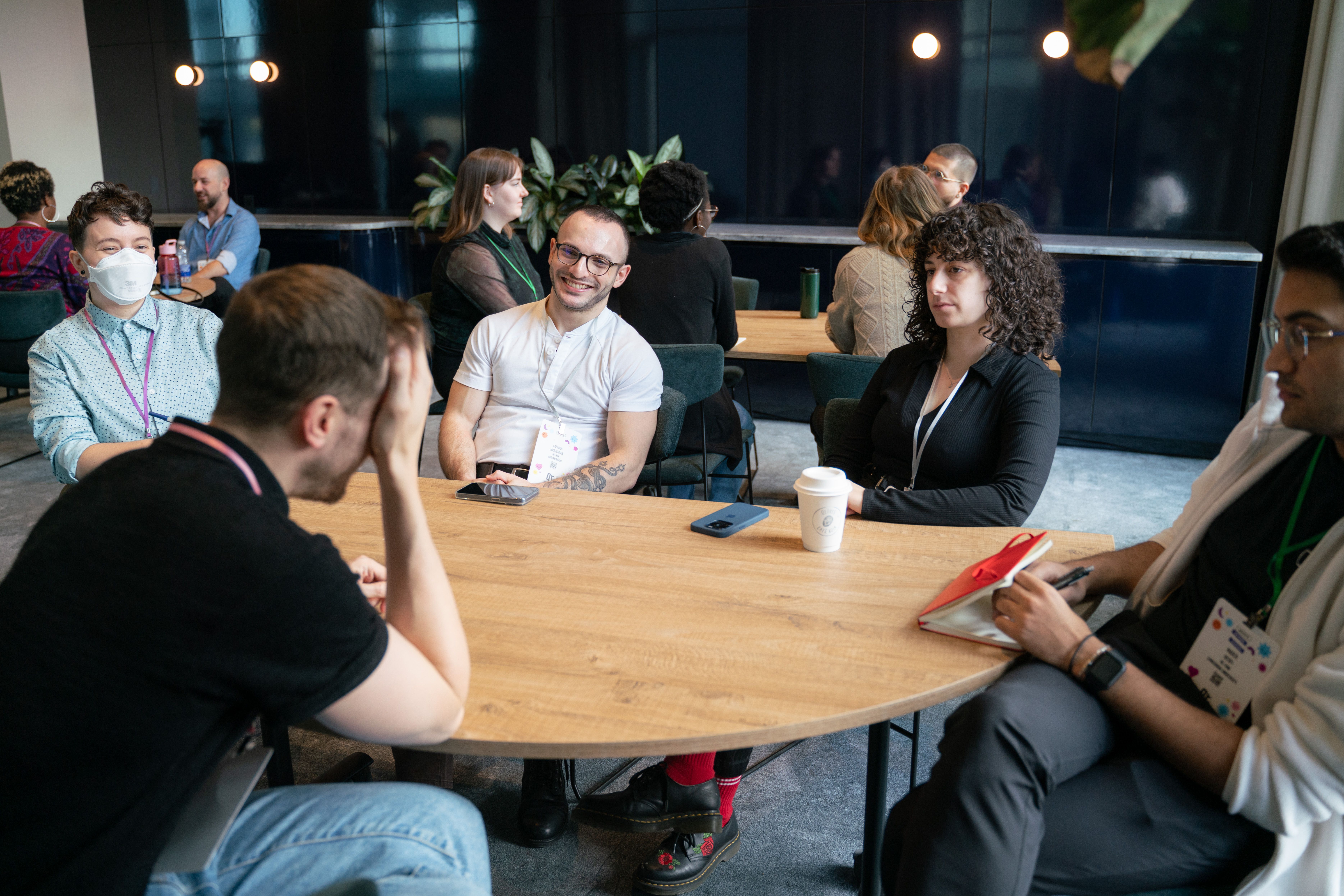 A group of people sit around a round wooden table in a modern conference room. One person wears a mask. The atmosphere appears casual and engaged.