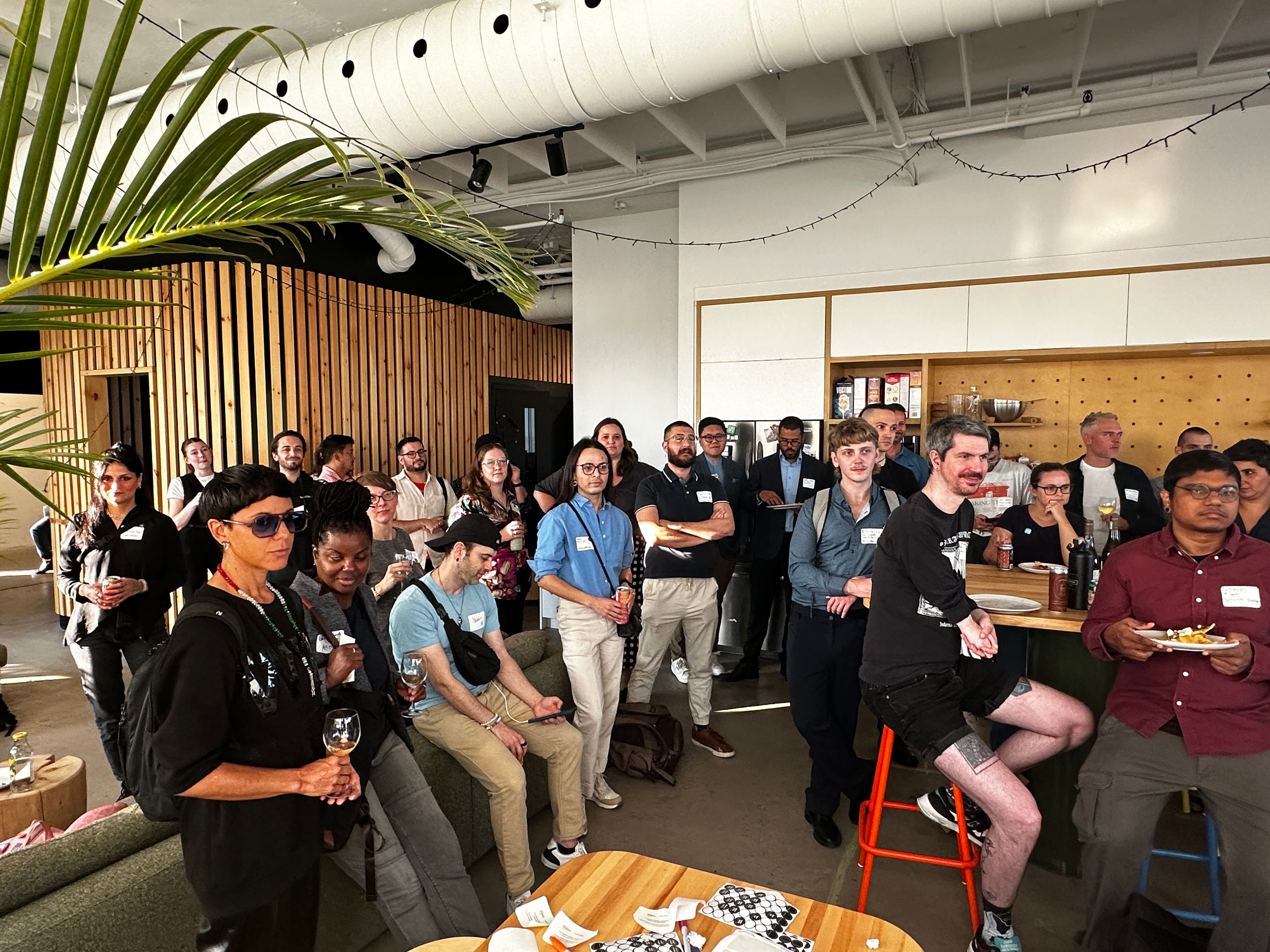 A diverse group of people, casually dressed, gather in a modern, well-lit room with wooden decor. They appear engaged, suggesting a relaxed social event. There are drinks and food, indicating a casual, friendly atmosphere.