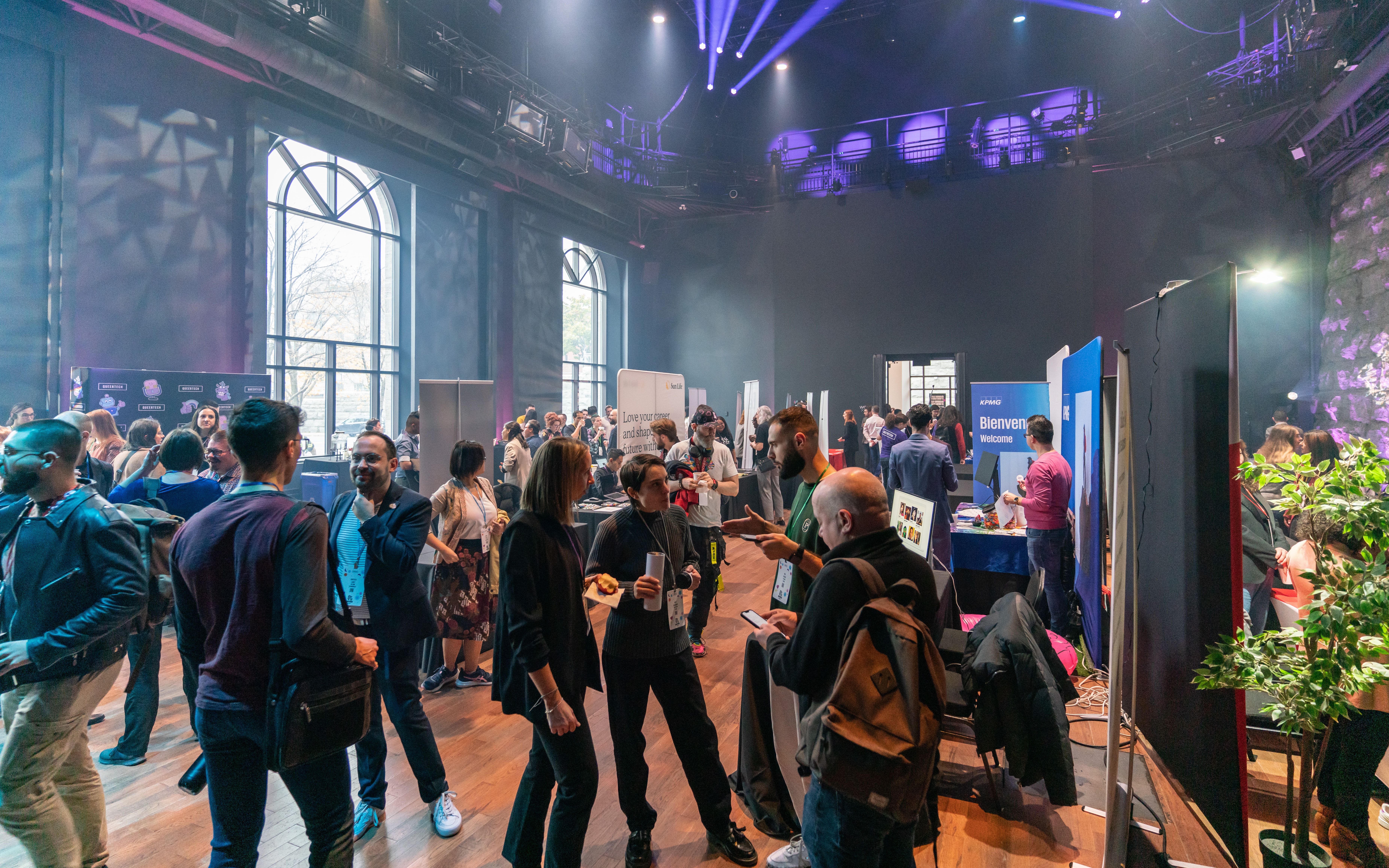 A large, dimly lit event space with people networking and lively conversations. Colorful booths line the walls, creating an engaging, vibrant atmosphere.