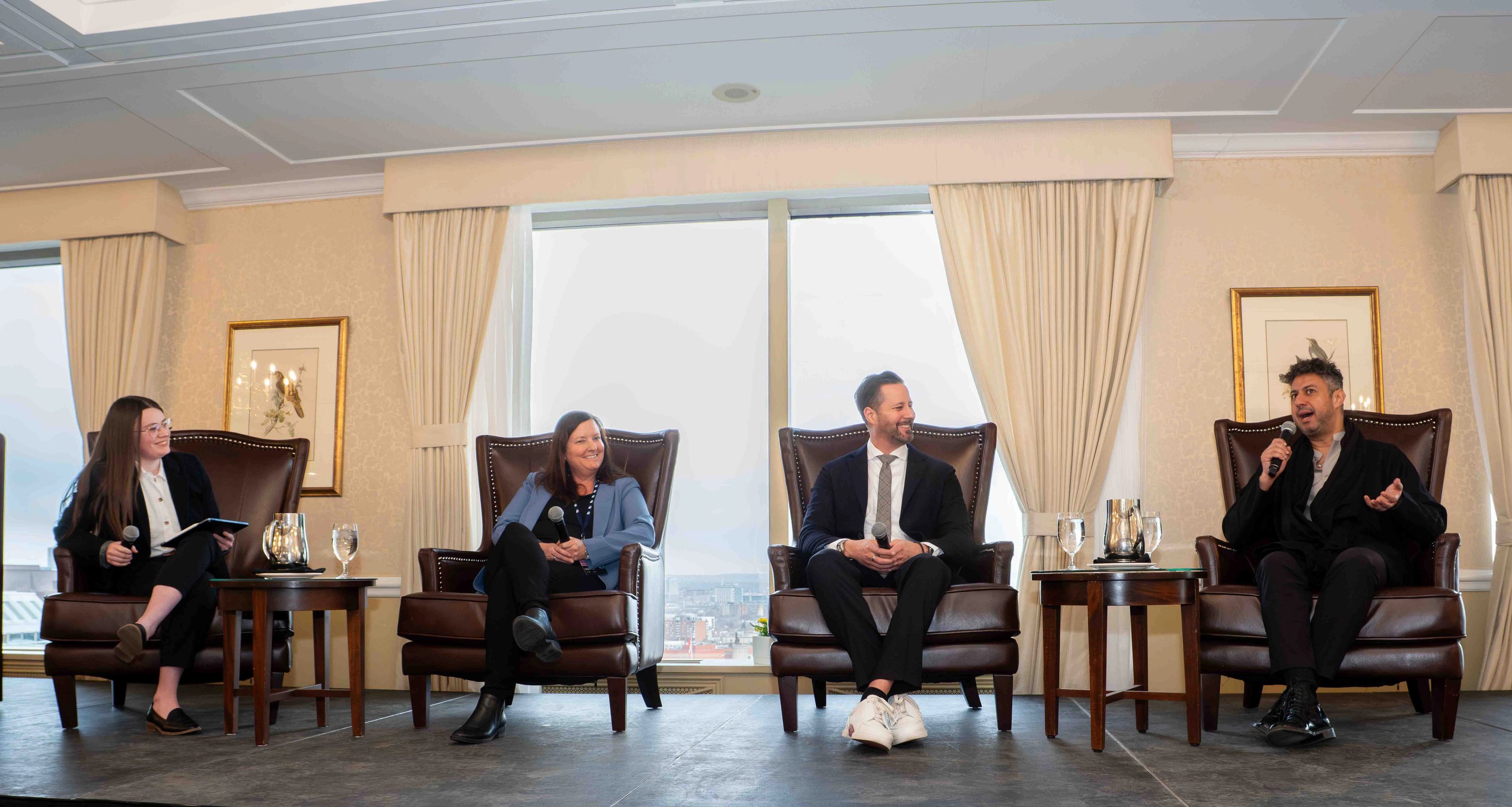 Panel discussion with four speakers seated on stage in armchairs, each holding a microphone, speaking in front of a large window backdrop.