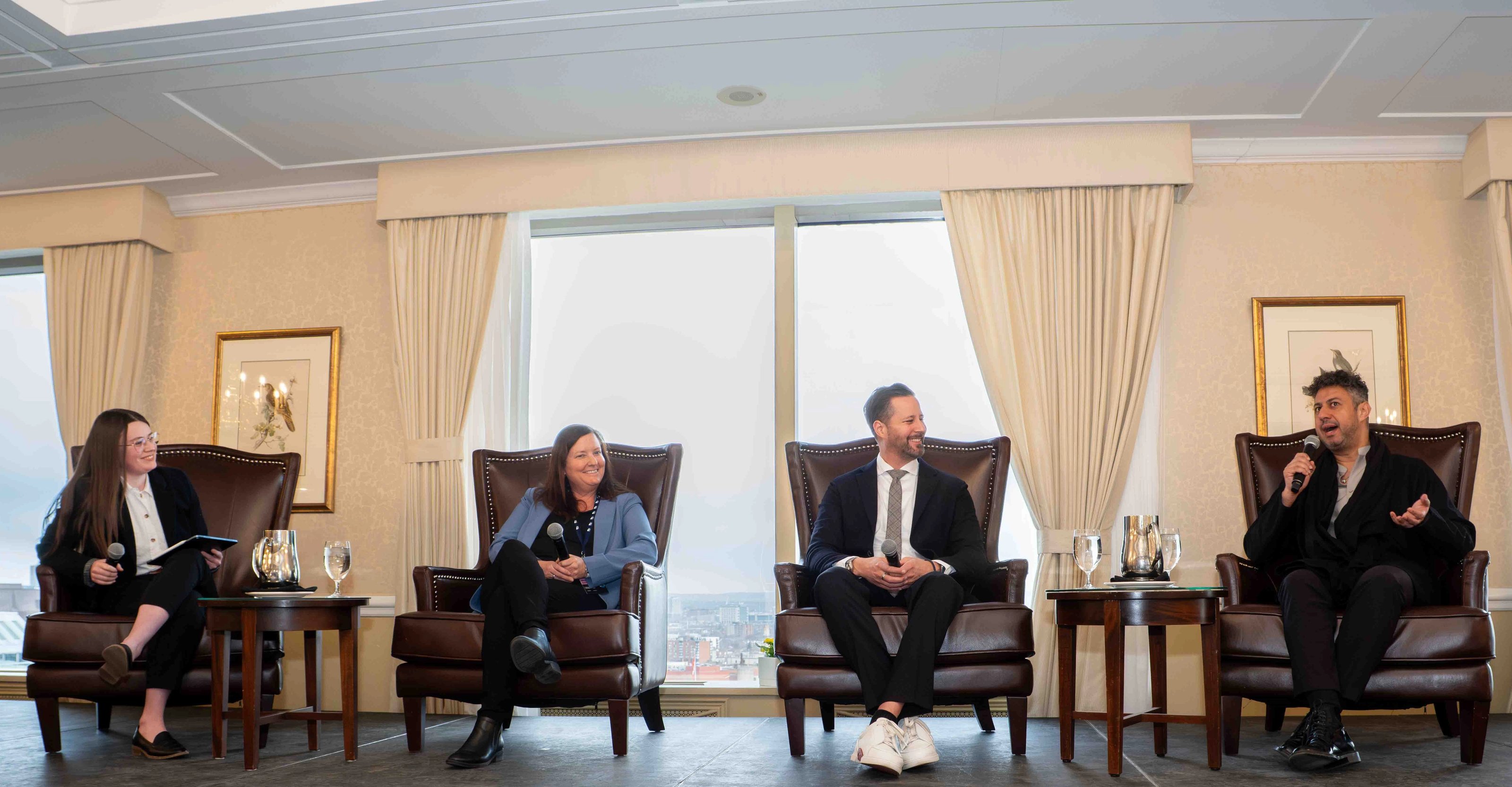 Discussion en panel avec quatre intervenant·e·s assis·es sur scène dans des fauteuils, chacun·e tenant un micro, devant un grand fond de fenêtres.