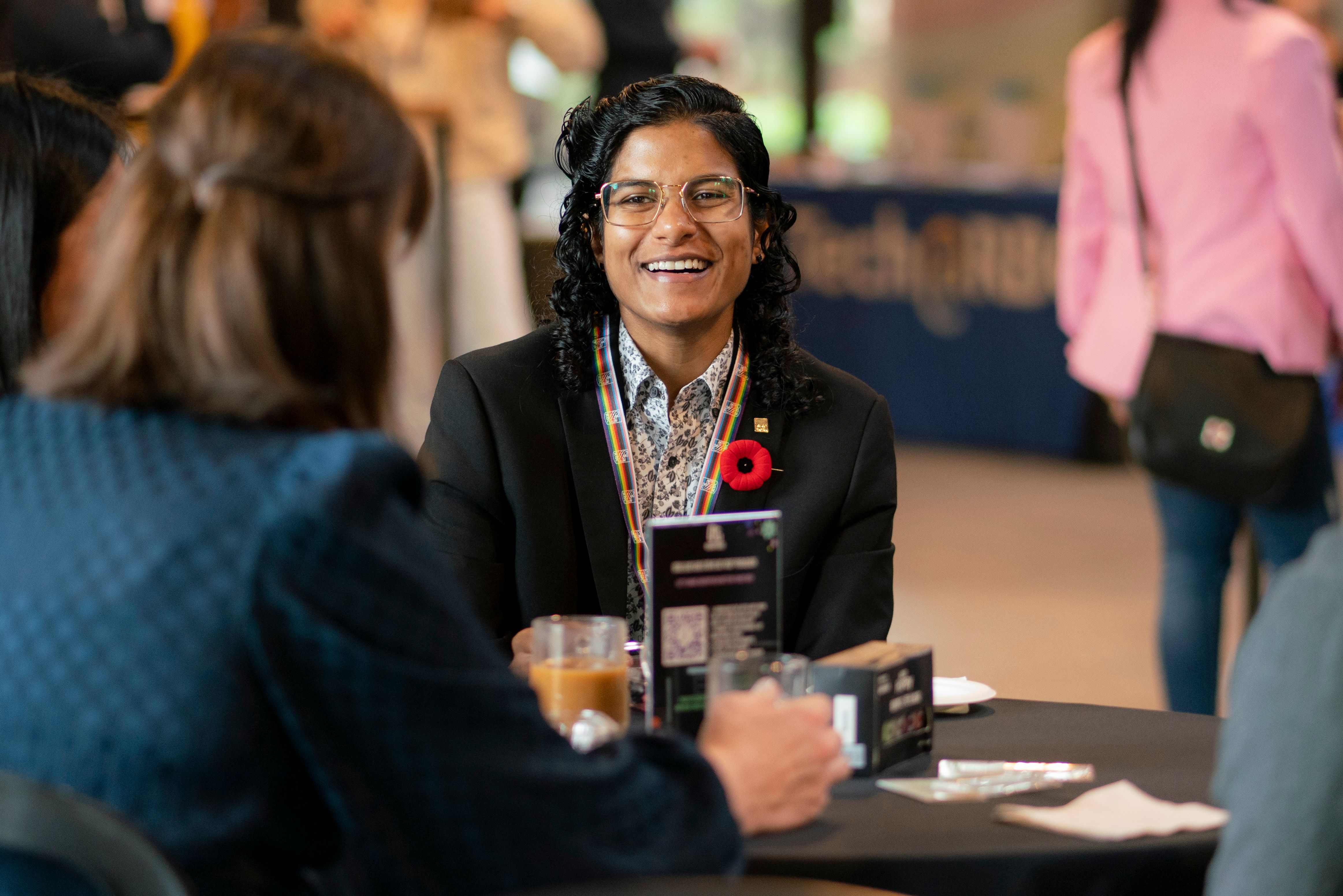 A smiling person with glasses, wearing a suit jacket and a poppy, sits at a table, engaging with others in a warm, positive atmosphere.