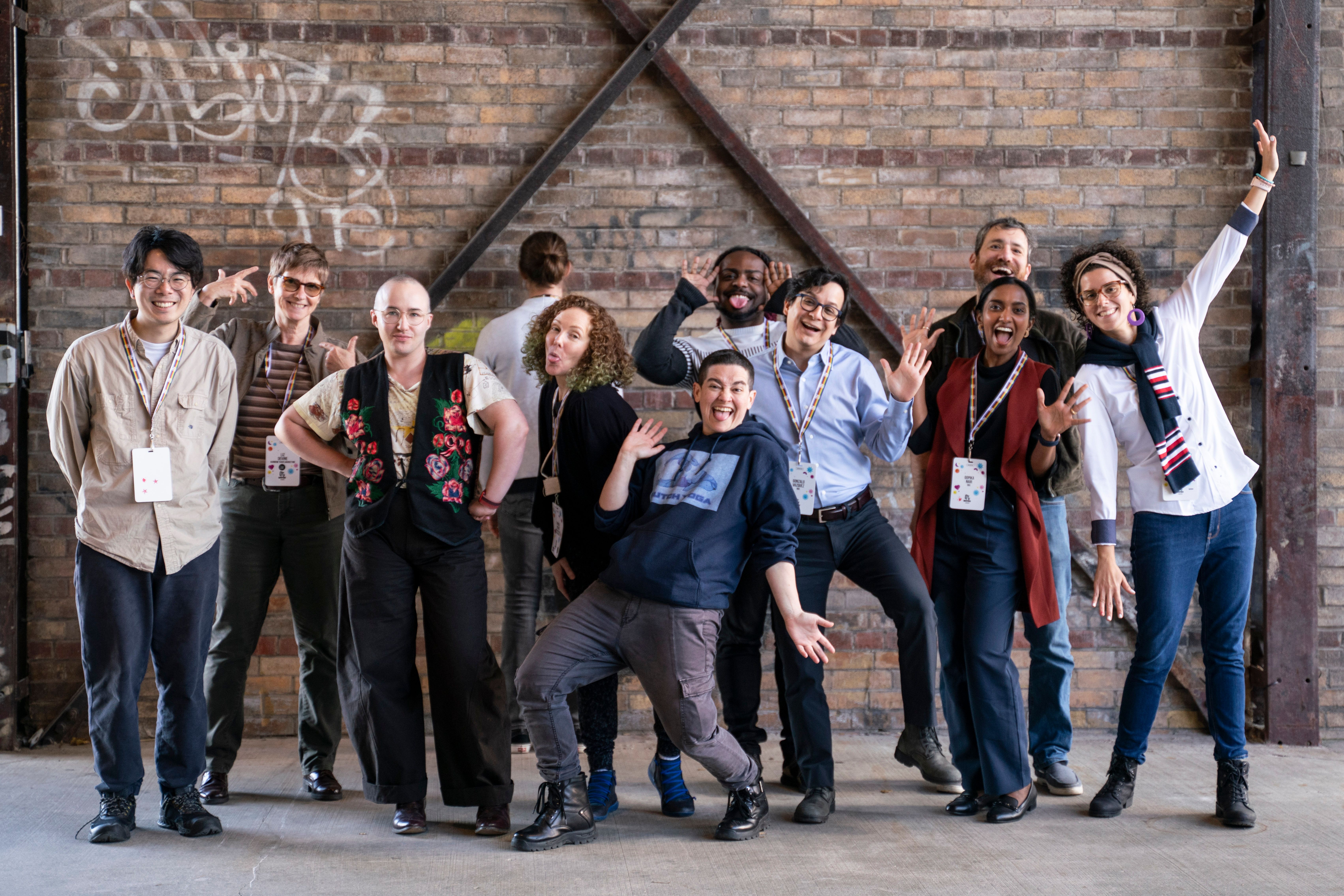 Groupe diversifié de participant·e·s posant de manière ludique devant un mur de briques, souriant et célébrant la communauté.