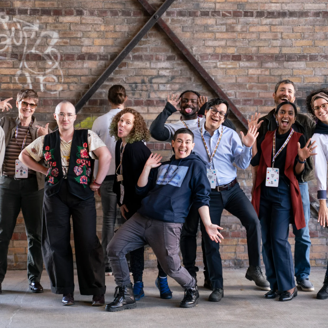 Groupe diversifié de participant·e·s posant de manière ludique devant un mur de briques, souriant et célébrant la communauté.