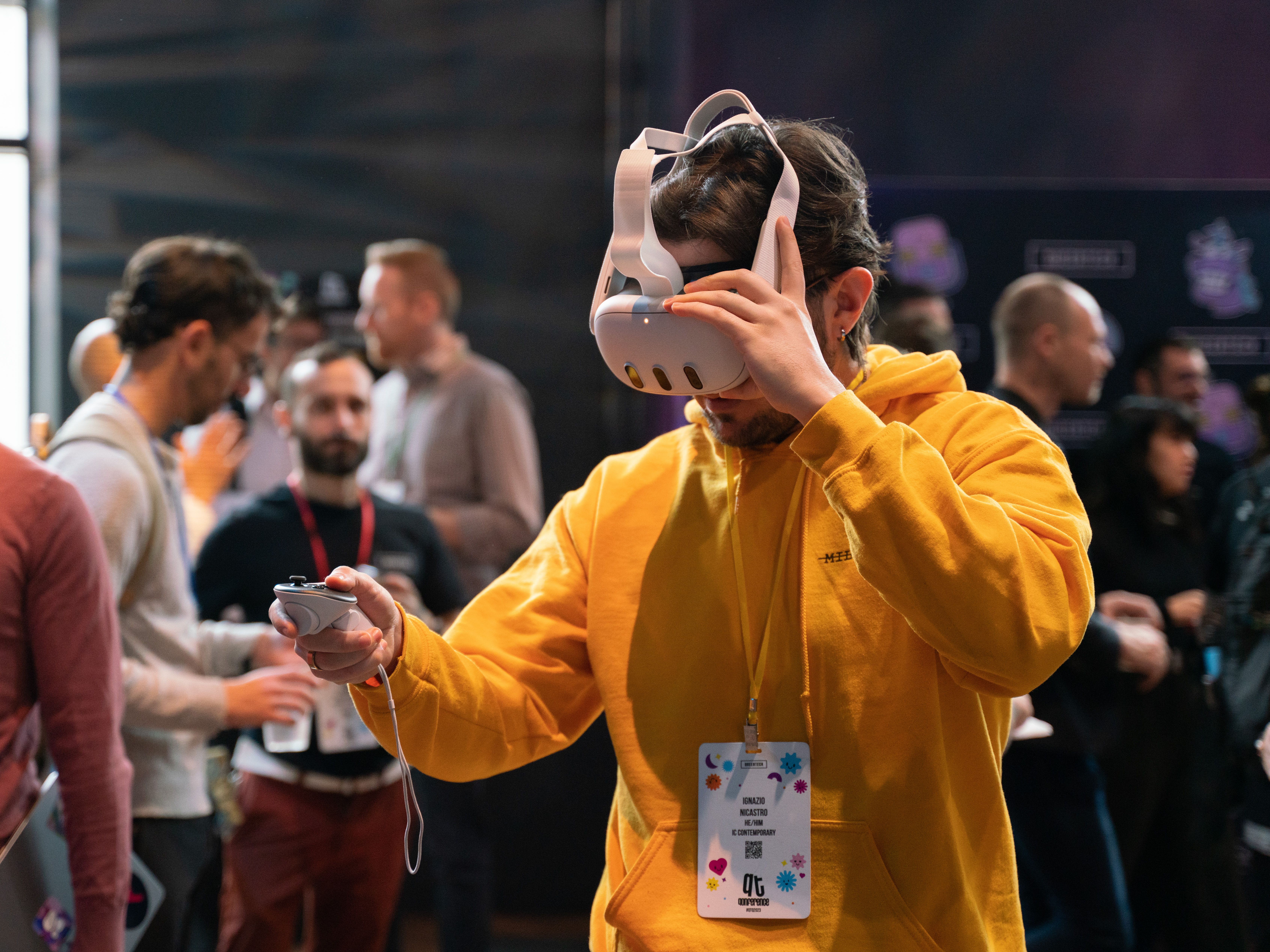 A man in a yellow hoodie uses a VR headset and controller at a tech event. A lanyard with a badge hangs from his neck. People chat in the background.