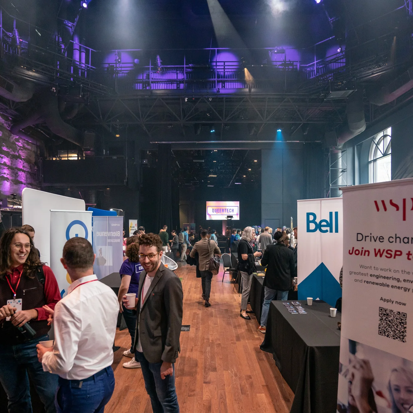 A bustling tech conference with people mingling beside company booths in a large, industrial venue. Purple lighting creates a vibrant atmosphere.