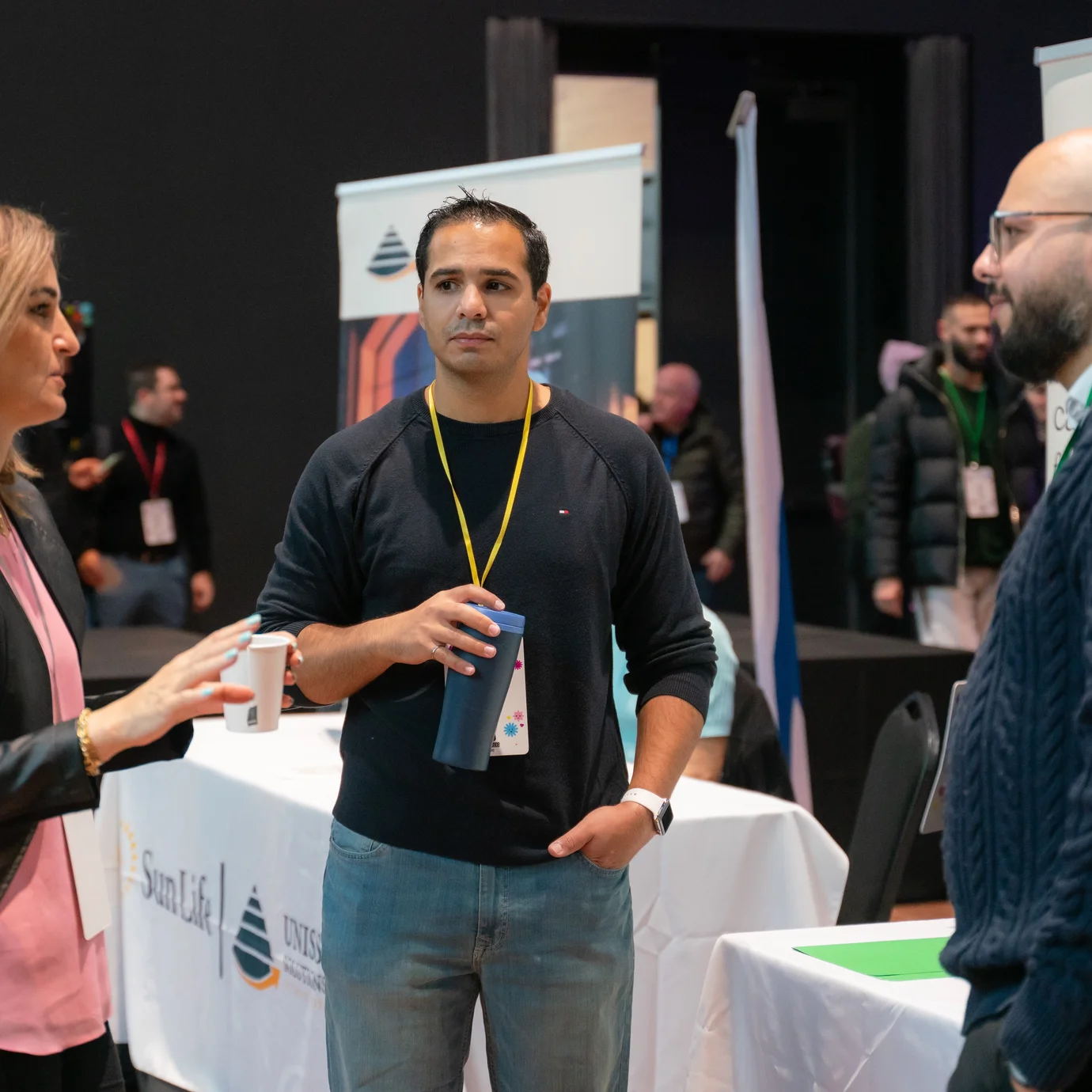 Three people converse at a networking event, surrounded by booths with company logos. The setting is professional, with a focused and engaged mood.