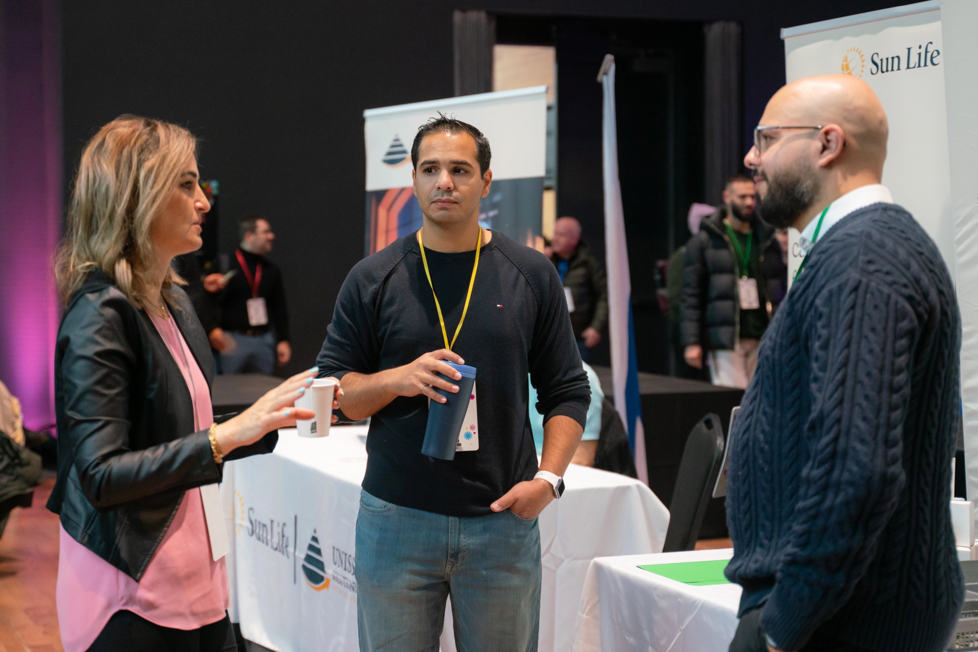 Three attendees standing and talking at a networking event beside sponsor booths, including a Sun Life display, while holding beverages and wearing event lanyards in a conference hall.