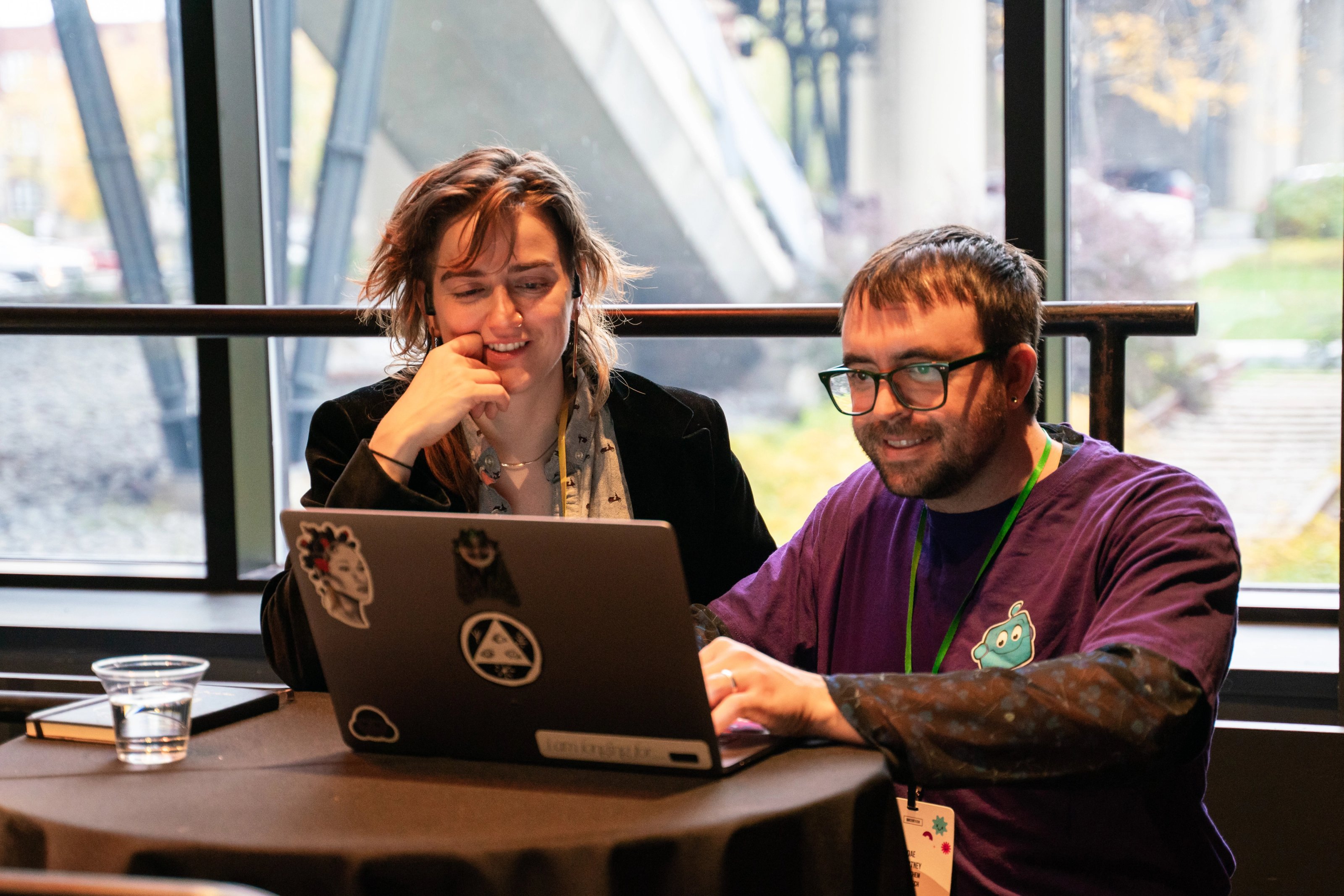 Two people smiling and looking at a laptop with stickers, seated at a round table with a notebook and glass. The room has large windows showing an outdoor scene.