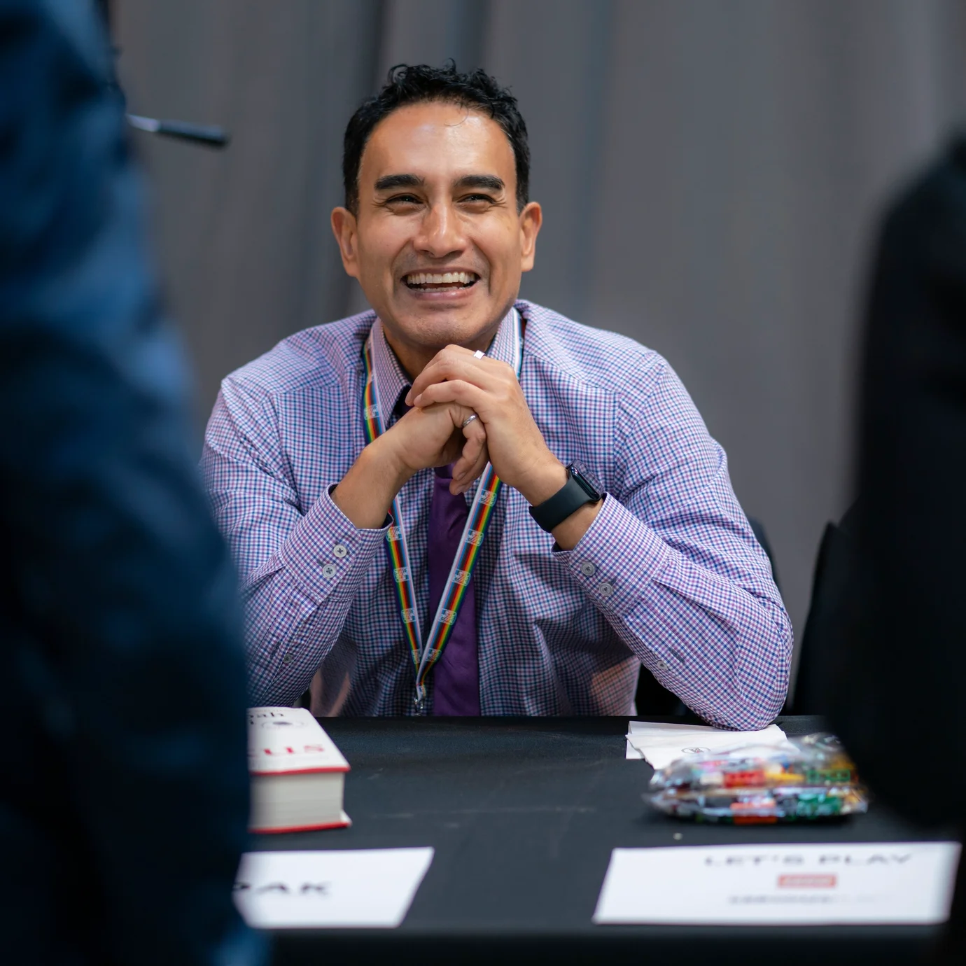 A man with dark hair smiles warmly, seated at a black table with a lanyard around his neck. The atmosphere is friendly and professional.