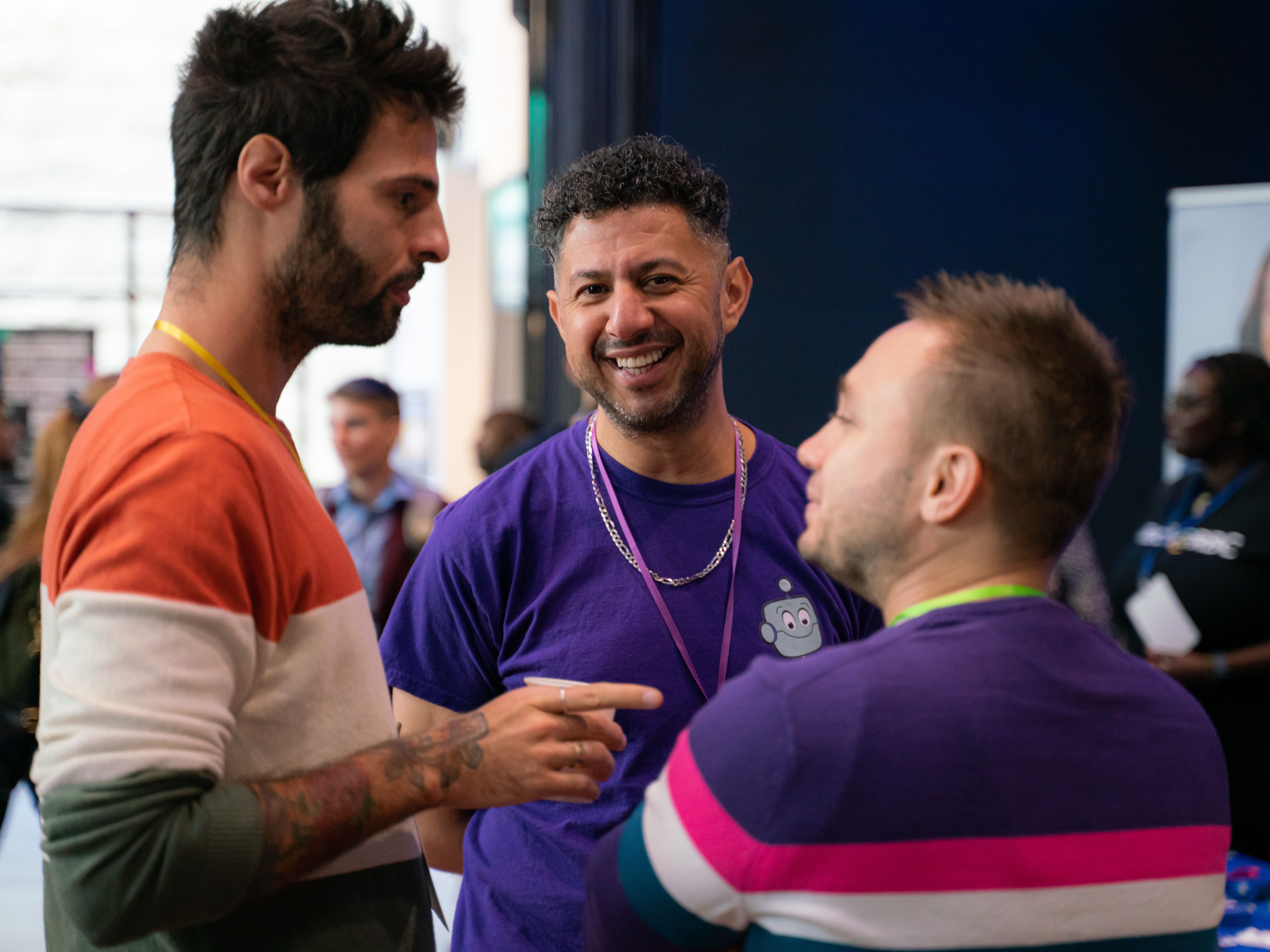 Three men engaging in a lively discussion. The man on the left wears a colorful sweater, the middle man a purple shirt, smiling, and the right man, a striped top. The atmosphere is friendly and casual.