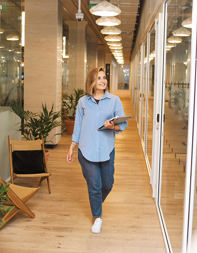 A woman in casual attire walks through a modern office hallway holding a laptop. She smiles, exuding confidence. Plants and warm lighting create a welcoming ambiance.