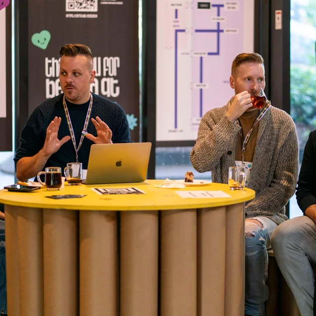 Four people sit around a yellow table having a discussion. One person gestures with their hands while another sips from a cup. Conference name tags hang around their necks, and posters are visible in the background. The atmosphere appears collaborative and informal.