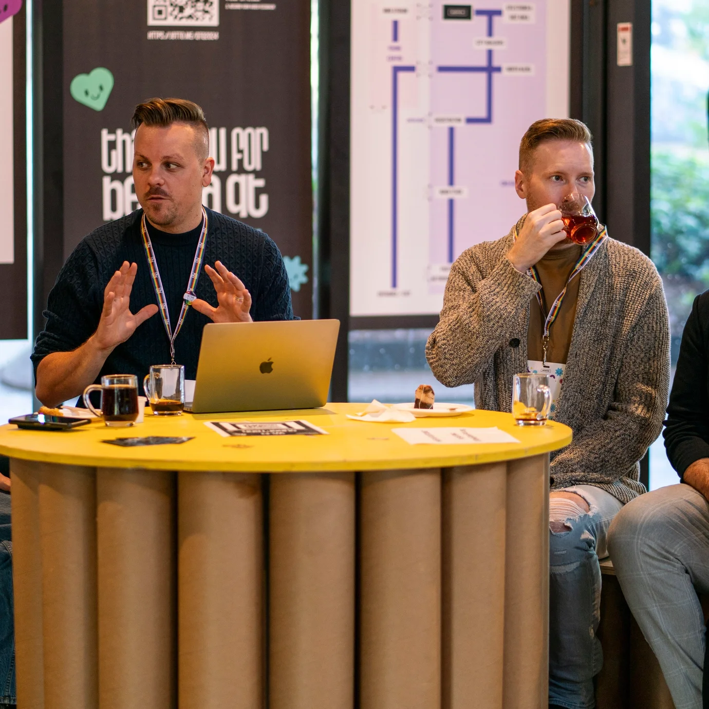 Four people sit around a yellow table having a discussion. One person gestures with their hands while another sips from a cup. Conference name tags hang around their necks, and posters are visible in the background. The atmosphere appears collaborative and informal.