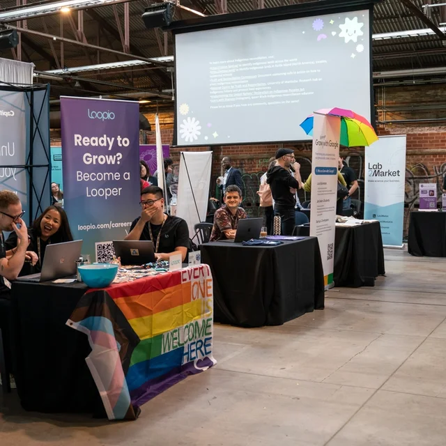 A vibrant career fair scene with people at booths. One booth is decorated with a rainbow flag, promoting inclusivity. The atmosphere is lively and welcoming.