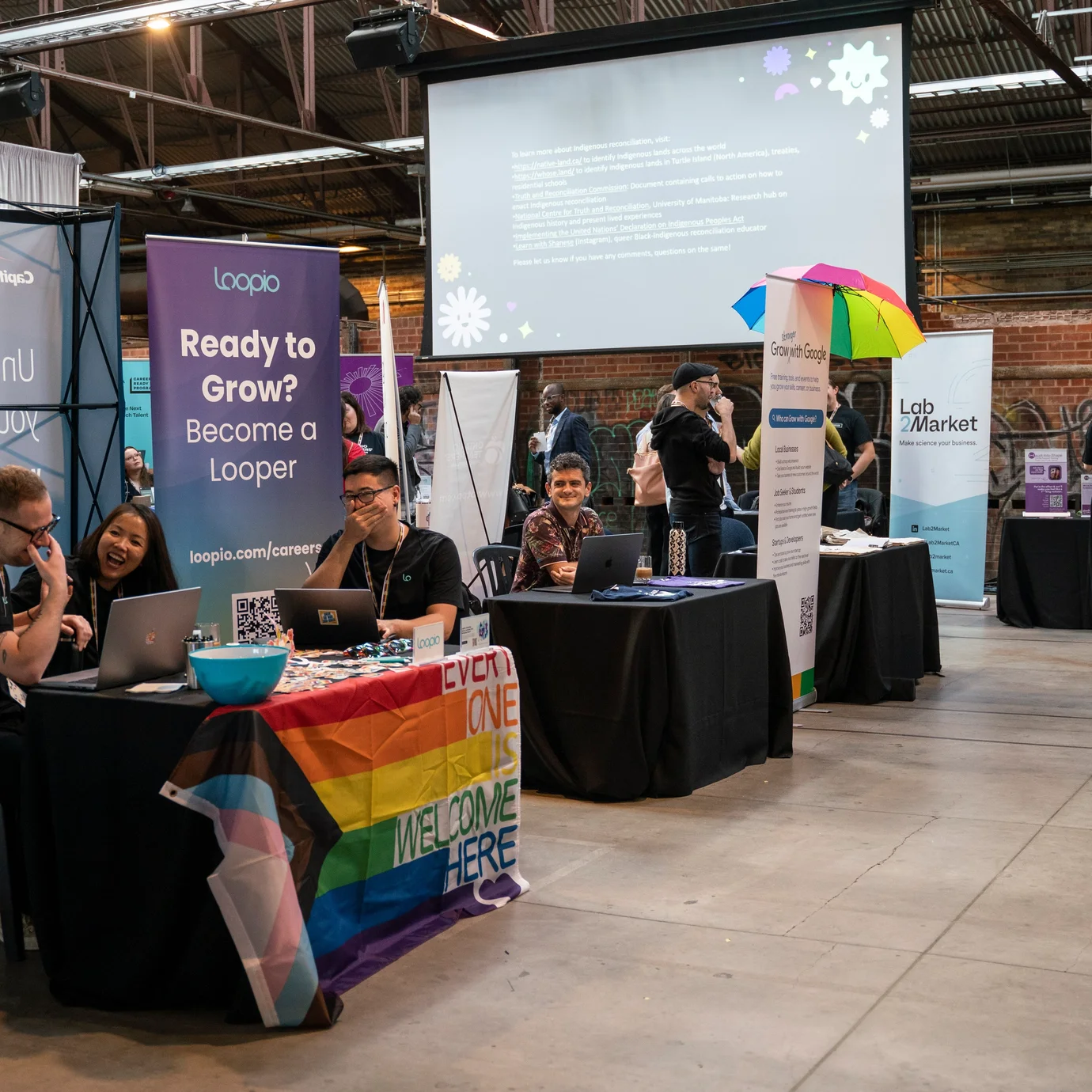 A vibrant career fair scene with people at booths. One booth is decorated with a rainbow flag, promoting inclusivity. The atmosphere is lively and welcoming.