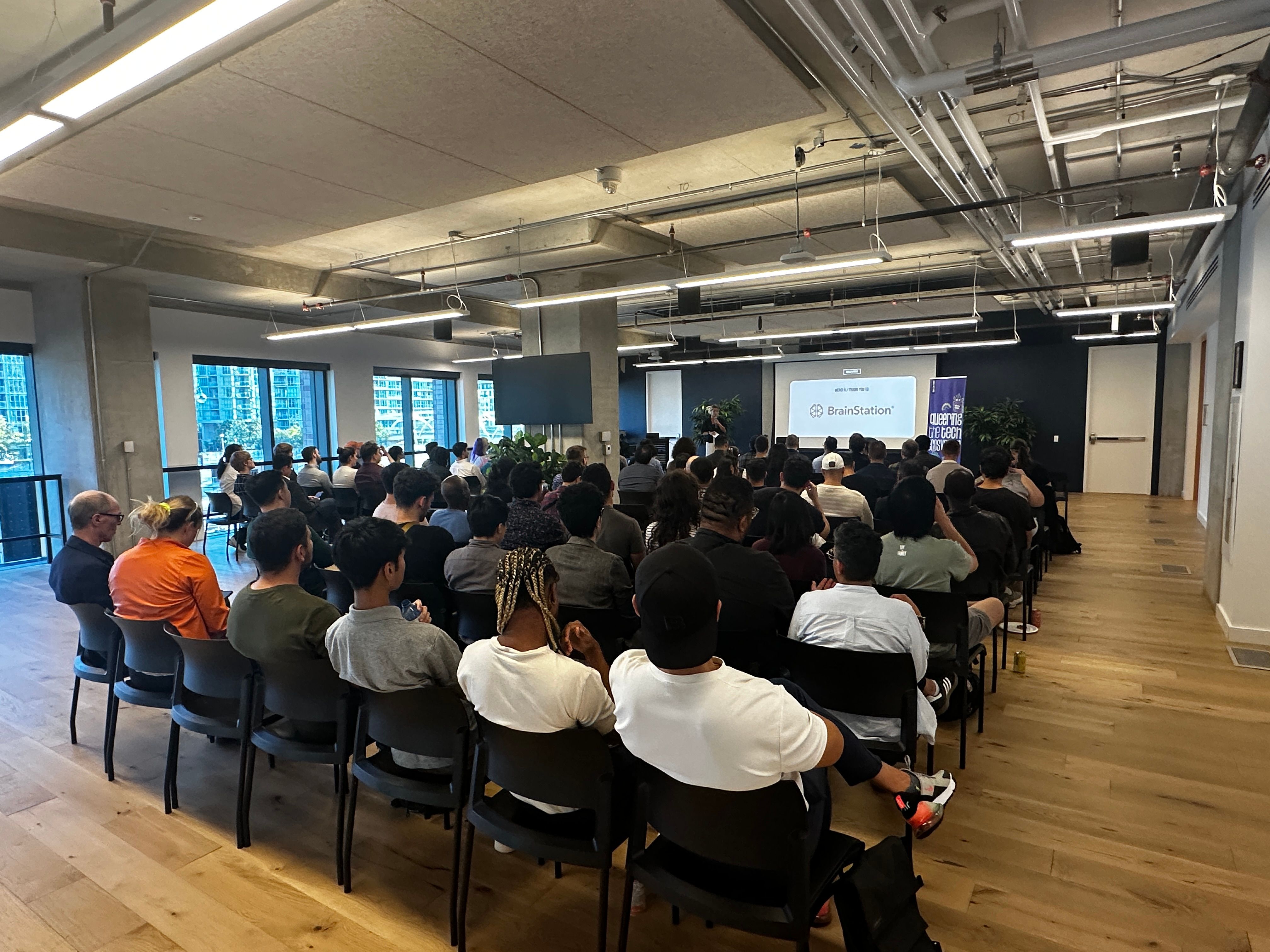 A large group of people sits in rows in a modern conference room with large windows. They face a presentation screen that displays a logo, suggesting a professional event or seminar.