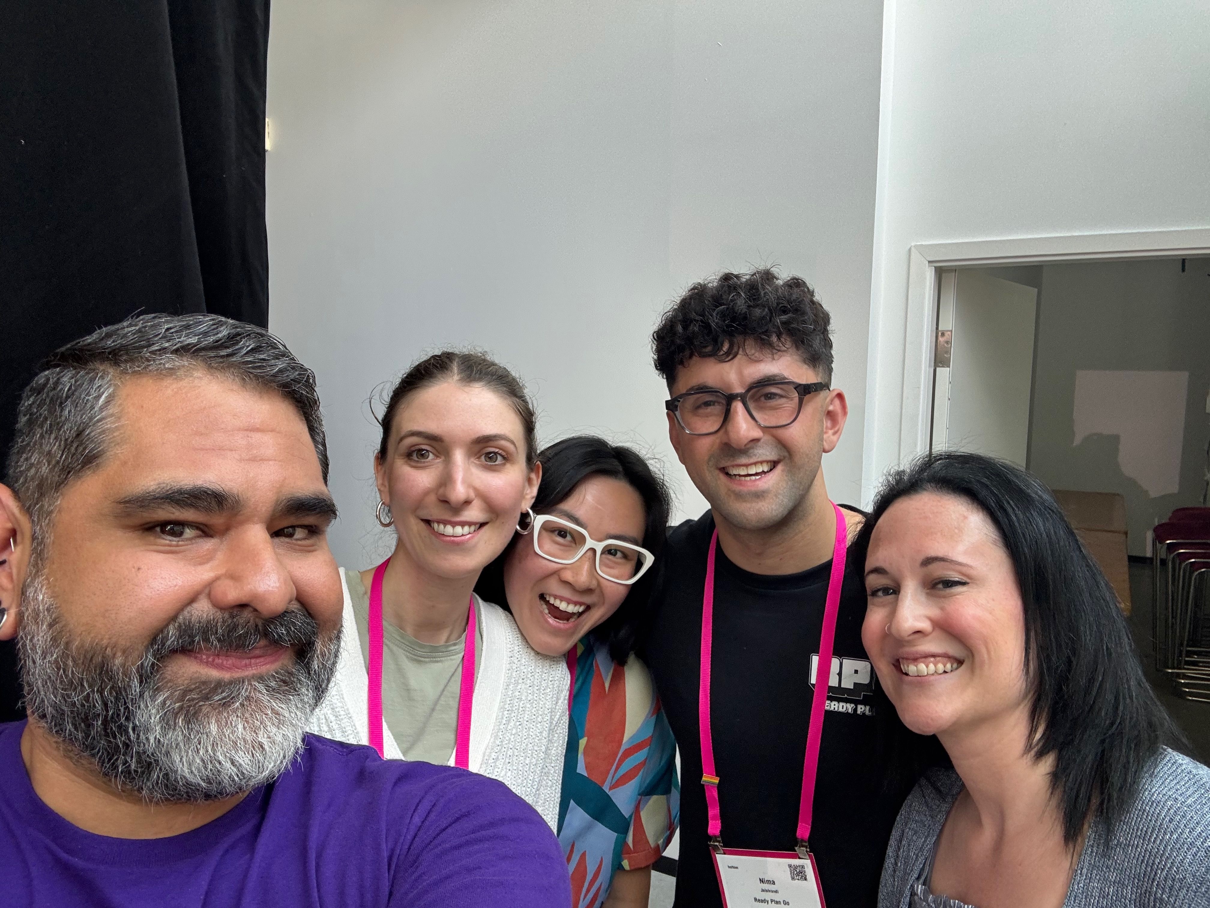 A group of five people smiling widely, standing close together indoors. They appear cheerful and friendly, suggesting a positive, social setting.