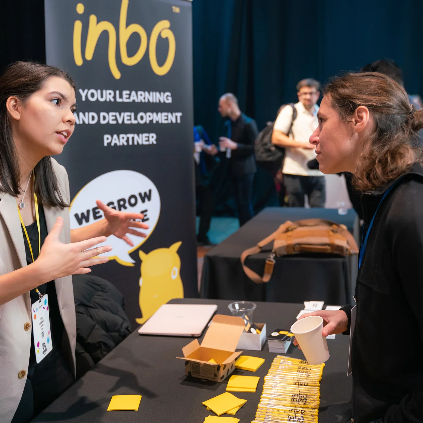 Two people engaged in a discussion at a conference booth with a "learning and development" banner, amid a busy, energetic networking event.
