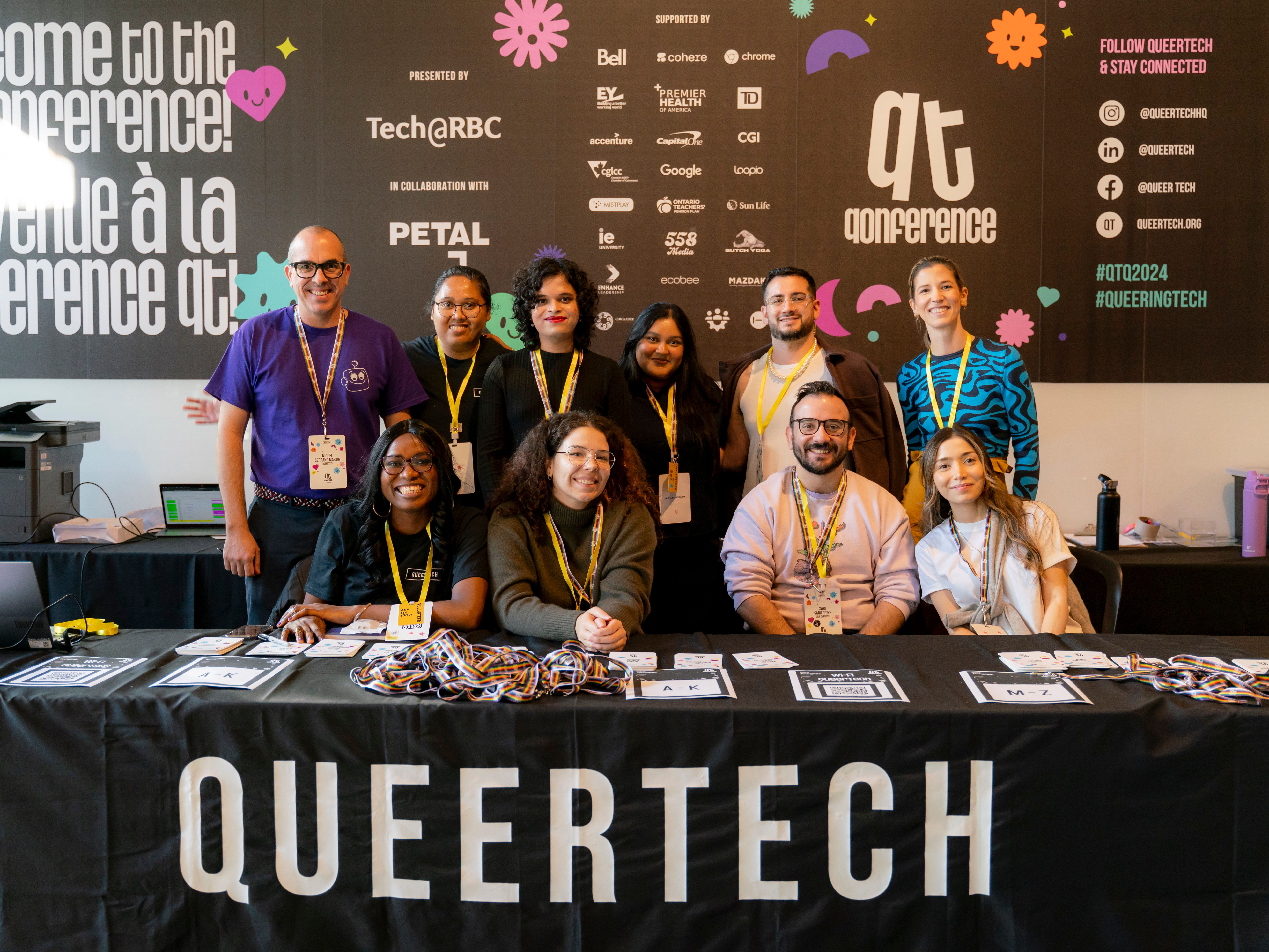 A group of ten people, smiling and diverse, pose behind a table with "QueerTech" banner at a tech conference. The backdrop is filled with logos and event branding.