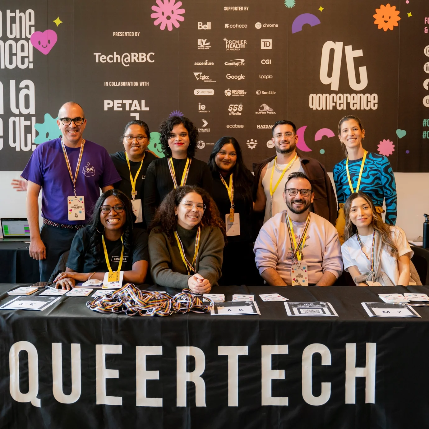 Groupe de participant·e·s de QTQ posant et souriant devant un mur de fond QueerTech dans un lieu de style industriel.