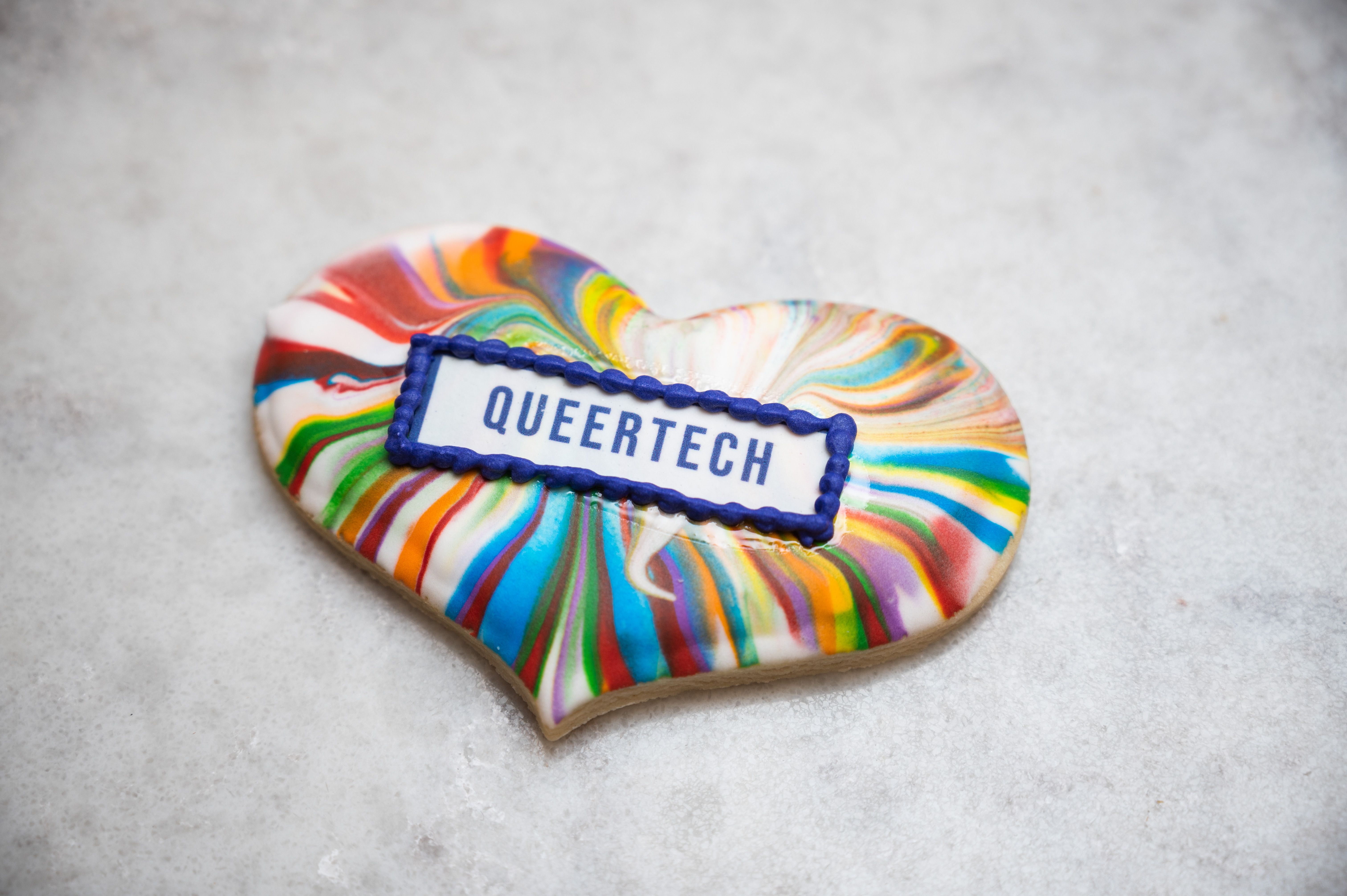 A heart-shaped cookie with a vibrant swirl of rainbow colors sits on a gray surface. The center features the word "QUEERTECH" in bold blue letters.
