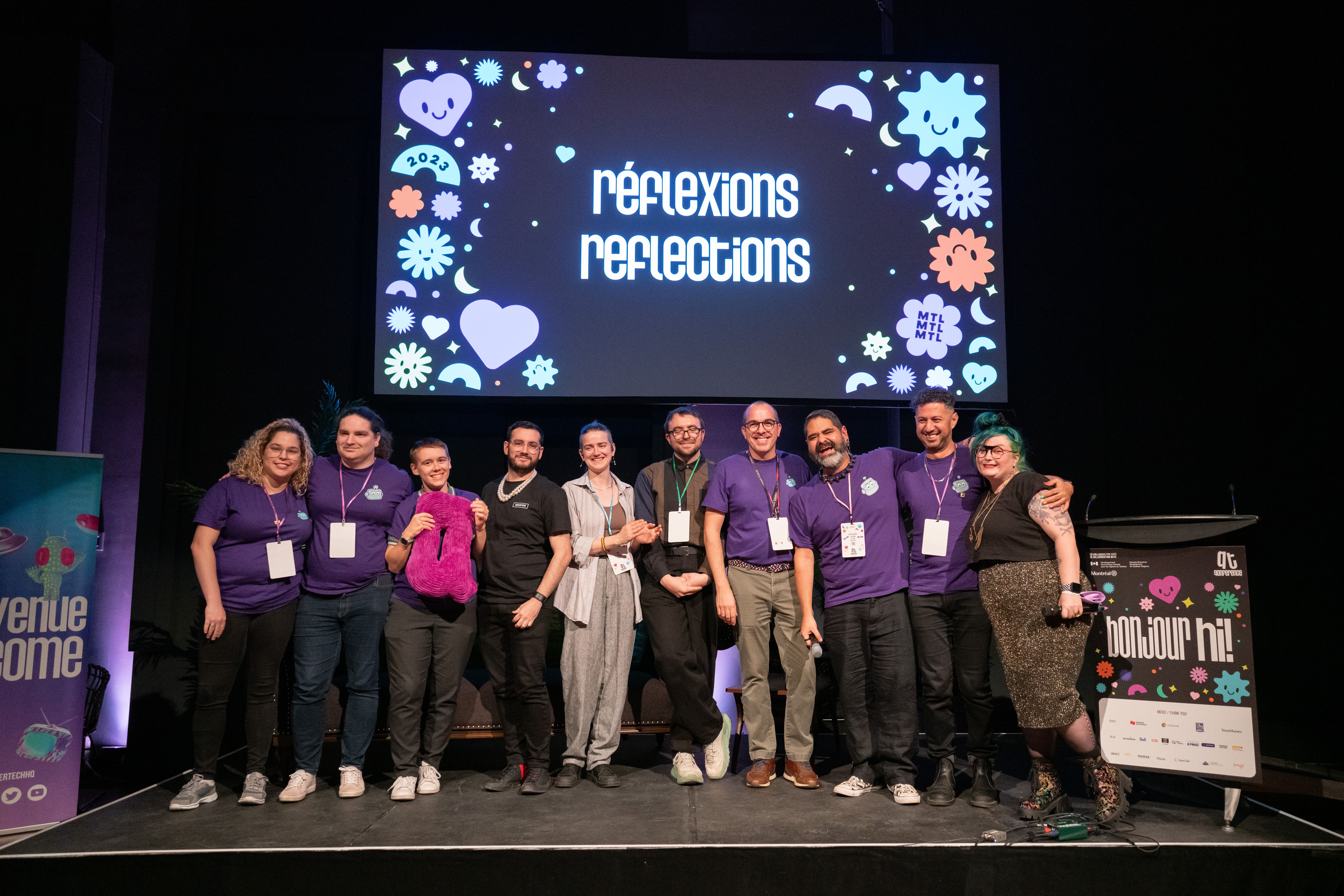 A group of smiling people stands on a stage. Behind them is a screen displaying "réflexions reflections". The atmosphere is friendly and welcoming.