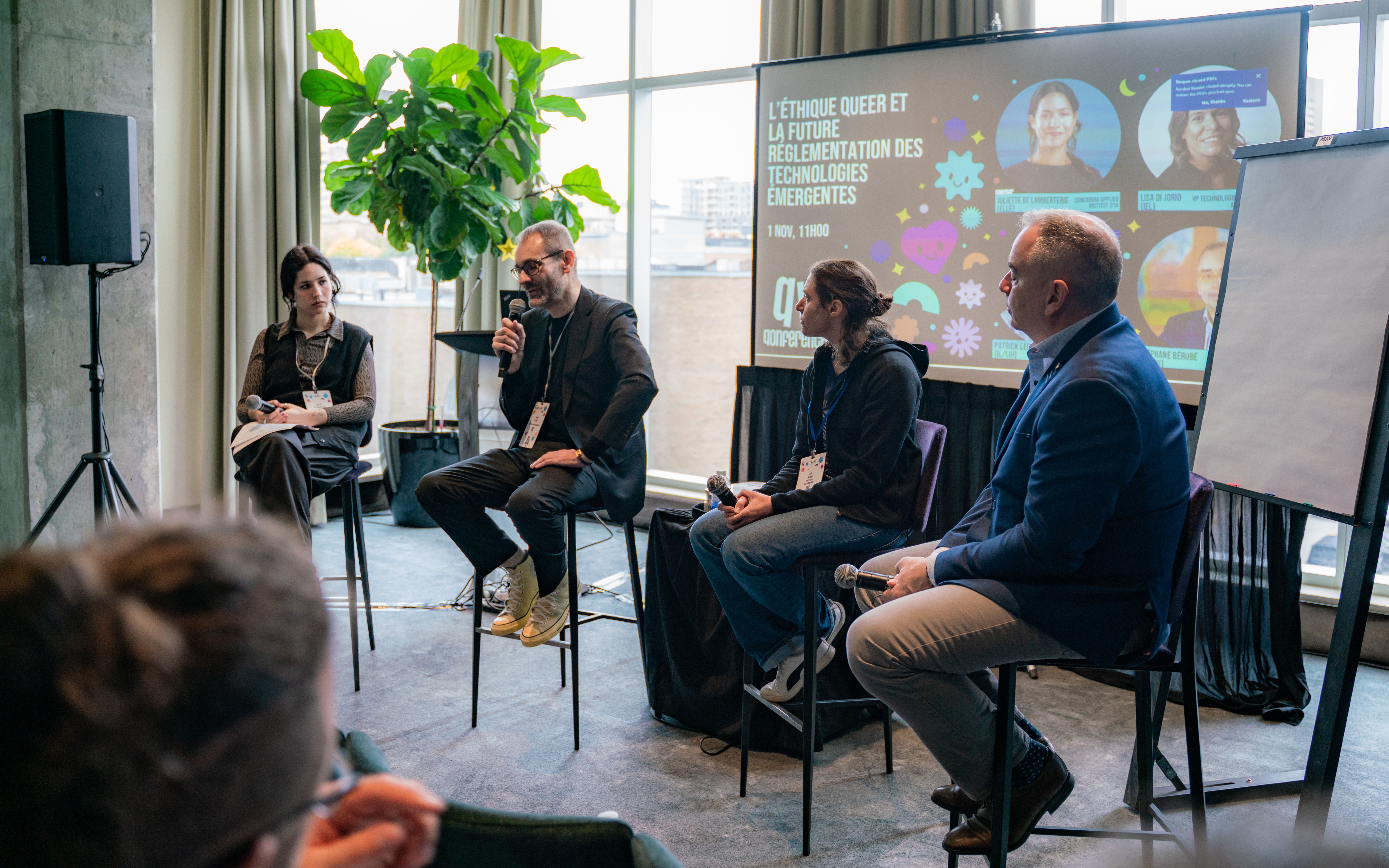 A panel of four people seated in a modern conference room. One person speaks into a microphone, engaging the audience. A projection screen displays discussion details on queer ethics and technology. The atmosphere is thoughtful and professional.