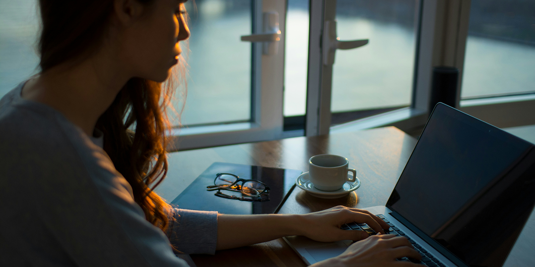 A woman types on a laptop in a dimly lit room with a large window showing dusk outside. A cup, tablet, and glasses lie nearby, creating a focused, serene atmosphere.