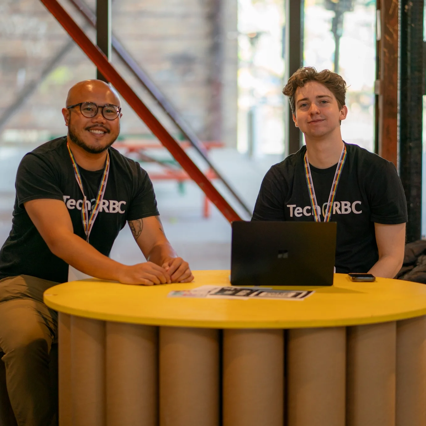 Two smiling people sit at a round, yellow table with a laptop, wearing Tech@RBC shirts. The setting is bright and casual, with a modern vibe.