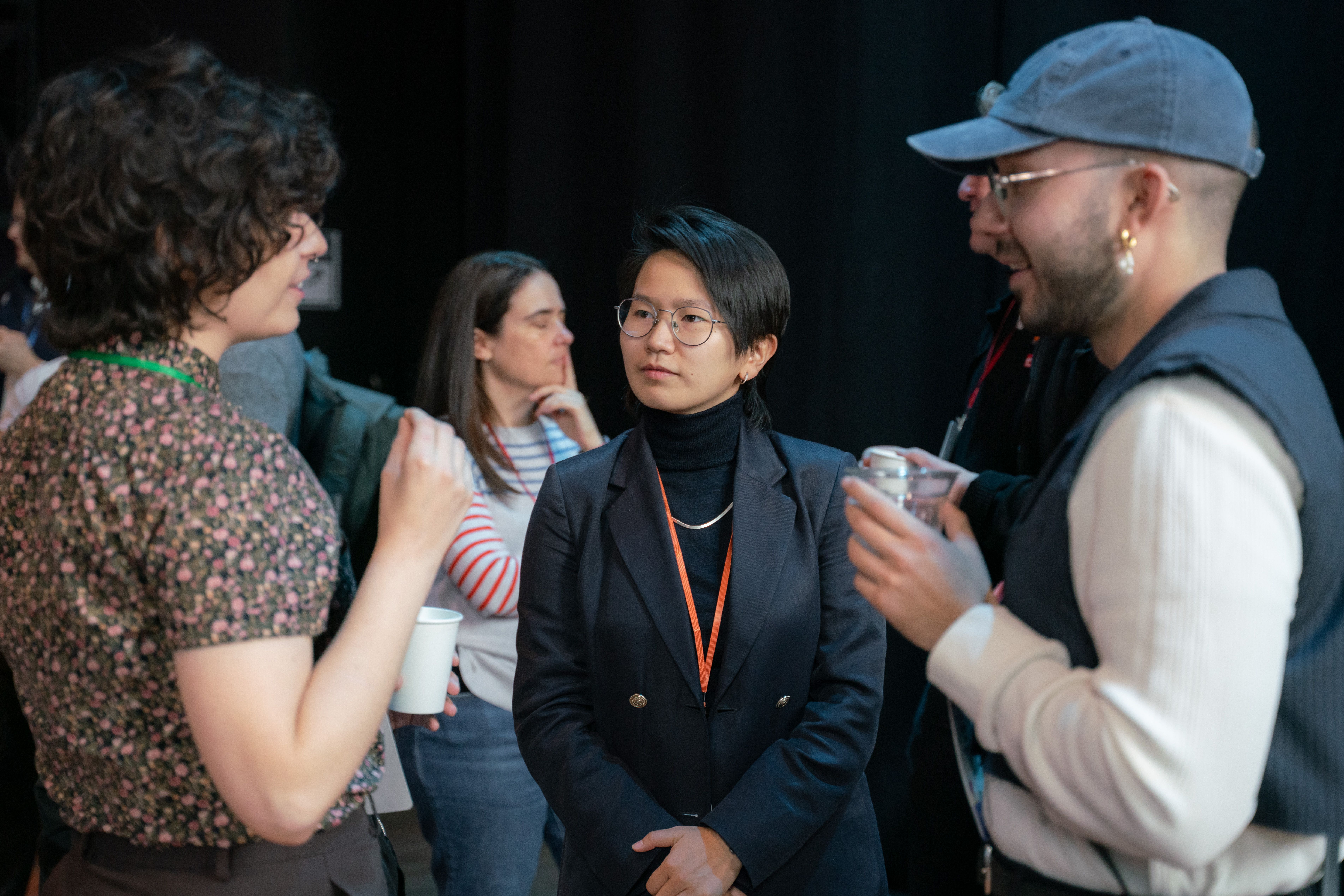 A group of people socializing at an event. A person in a blazer and glasses listens intently, while others engage in animated conversation. The atmosphere is lively.