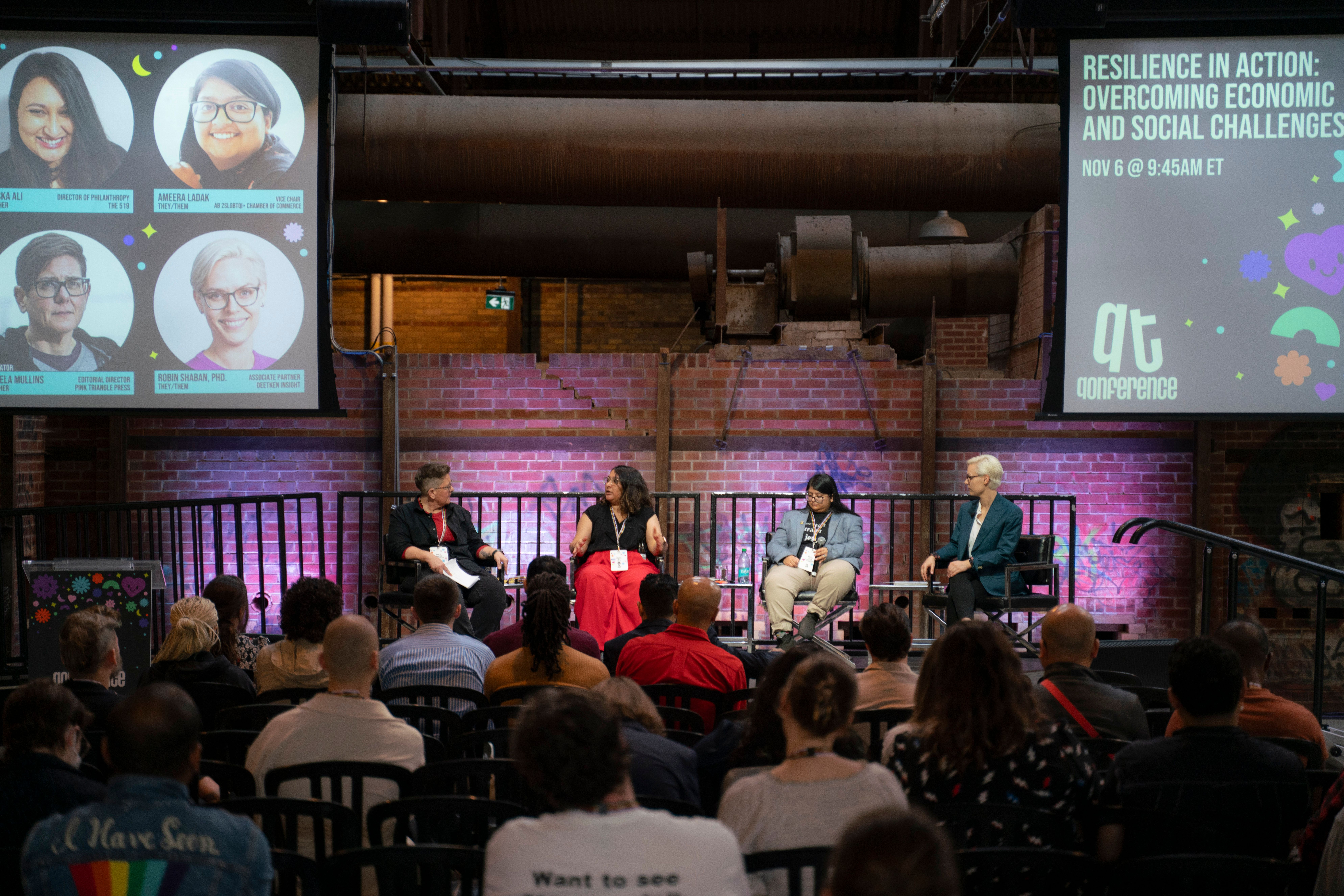 Panel discussion on stage at QTQ with speakers seated and audience watching, large screens displaying session details.