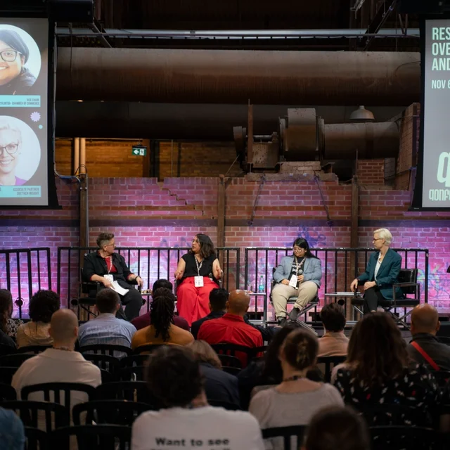 Panel discussion on stage at QTQ with speakers seated and audience watching, large screens displaying session details.