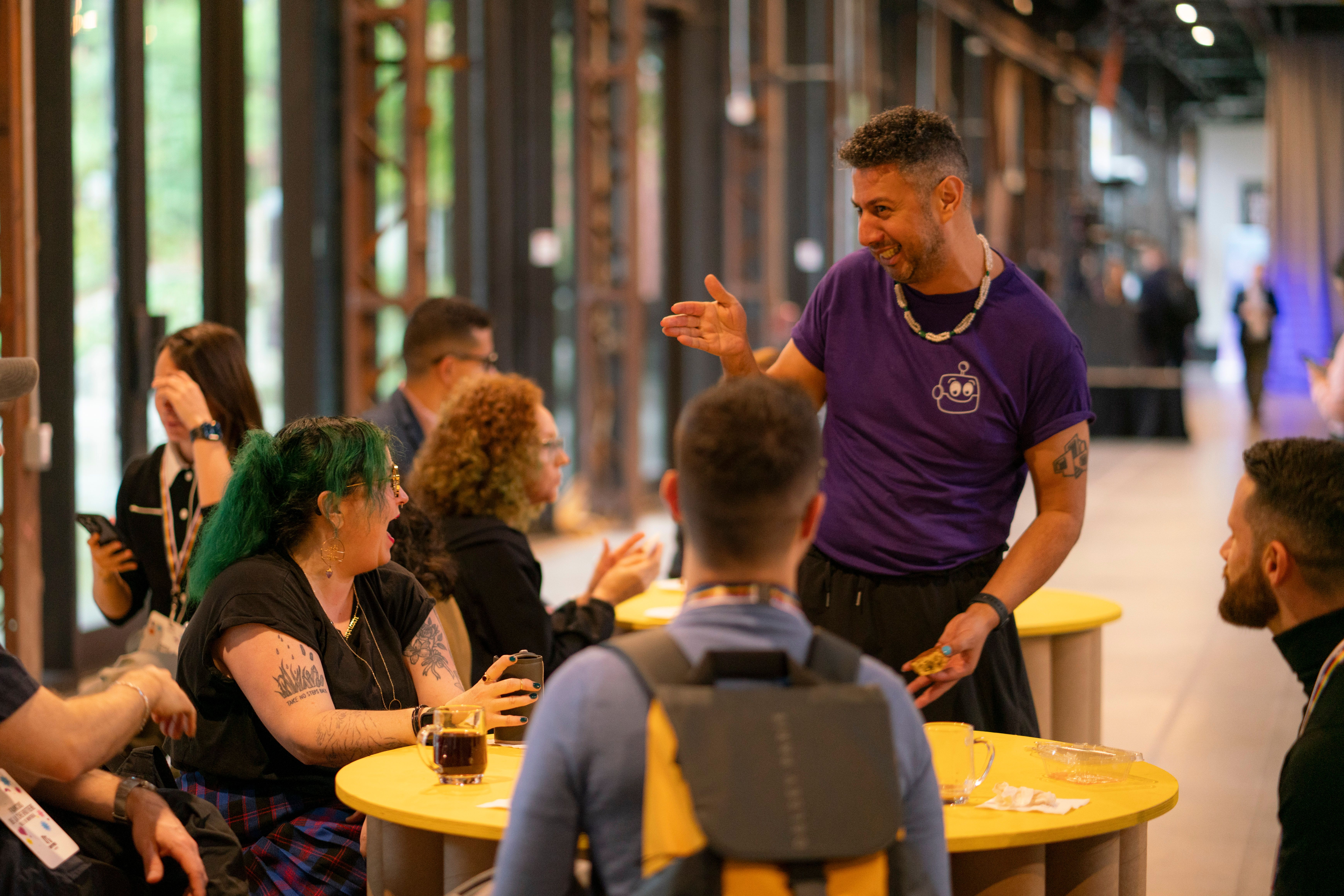 A man in a purple shirt animatedly speaks to a group seated at a yellow table indoors. People are engaged and smiling, creating a lively atmosphere.