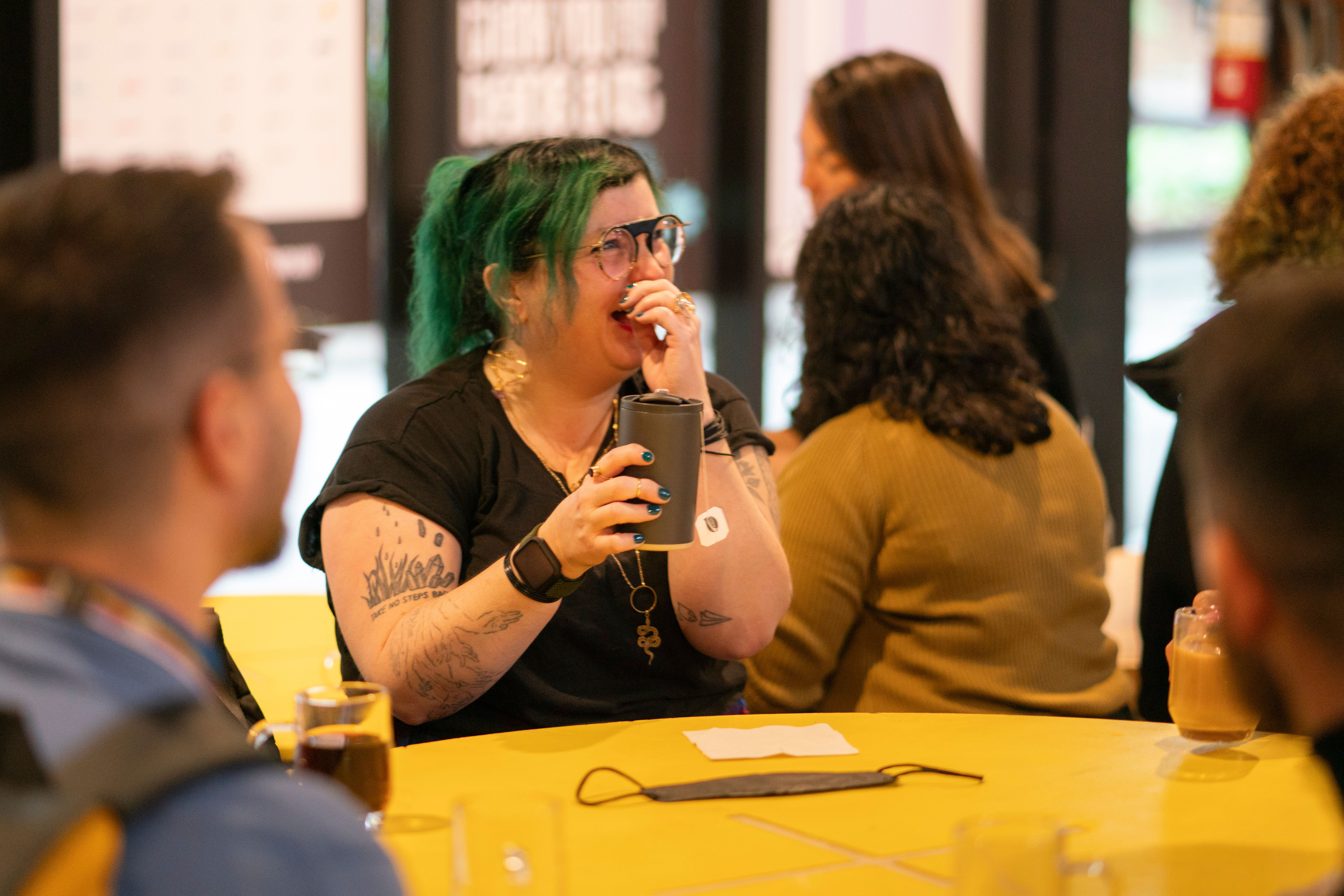 A person with green hair and tattoos laughs while covering their mouth, holding a cup at a yellow table. Several others are sitting nearby, creating a warm, joyful atmosphere.