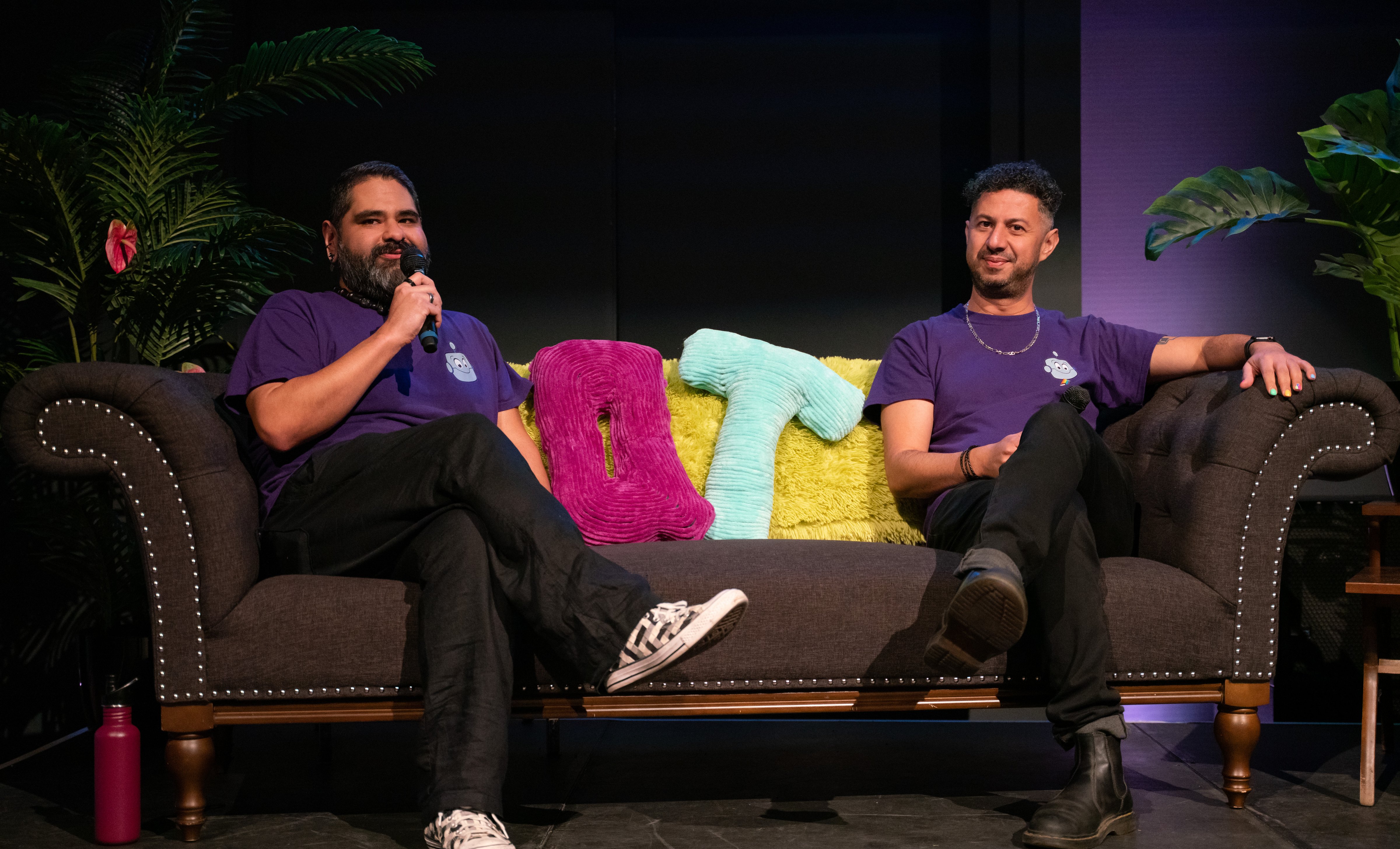 Two men in purple shirts sit on a brown couch with fluffy letters behind them. They appear relaxed, speaking into microphones with plants around.