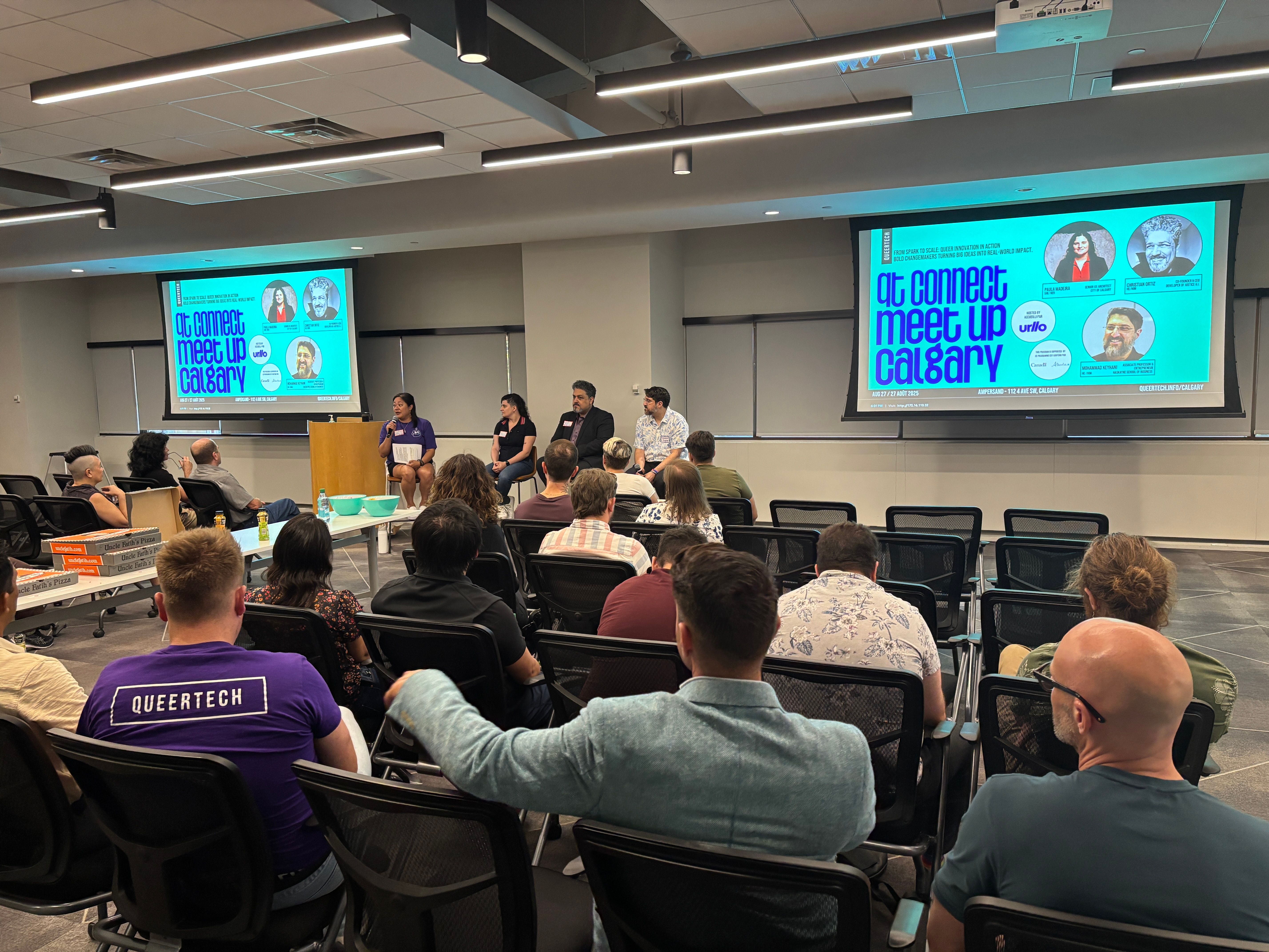 A group of people sit facing a panel of four speakers in an indoor meeting room. A large screen displays "qt connect meetup Calgary" with speaker photos. The audience appears attentive.